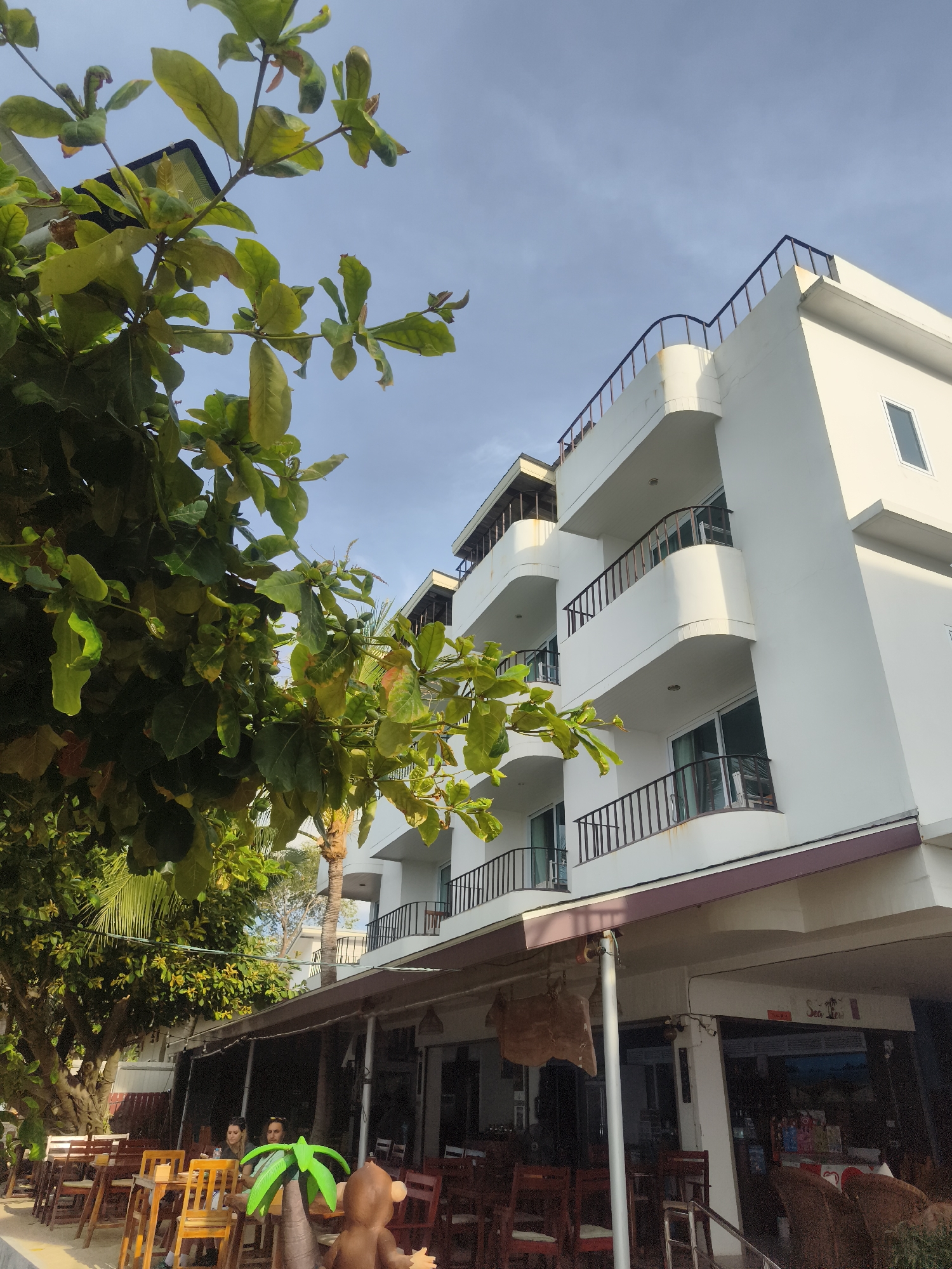 This image depicts a vibrant outdoor scene in Baan Mae Nam, Thailand, centered around a multi-story white building that appears to be a hotel or guesthouse, and an adjacent open-air restaurant.
The foreground and left side are dominated by lush, dense green foliage from large trees, their leaves illuminated by bright daylight, suggesting late morning or early afternoon. A clear blue sky with minimal cloud cover is visible above, indicating pleasant weather.
The main building is characterized by its clean white facade and multiple levels, each featuring rounded balconies with dark iron railings. A rooftop area, also with railings, is visible at the very top. The architecture is modern and functional.
At the ground level, beneath the building's overhang and an extended awning, is an outdoor dining area. Wooden tables and chairs, some with red cushioned seats, are neatly arranged for patrons. Two individuals, a man and a woman, are seated at one of these tables, engaged in conversation or relaxation. On a nearby table in the immediate foreground, a playful brown monkey figurine with a green palm tree on its head serves as a decorative element. Under the awning, a sign with the faintly visible text "Sea Life" is present.
The scene conveys a relaxed, tropical atmosphere, typical of a resort or coastal town. The bright sunlight, the presence of diners, and the surrounding verdant greenery all contribute to a feeling of leisure and vacation.