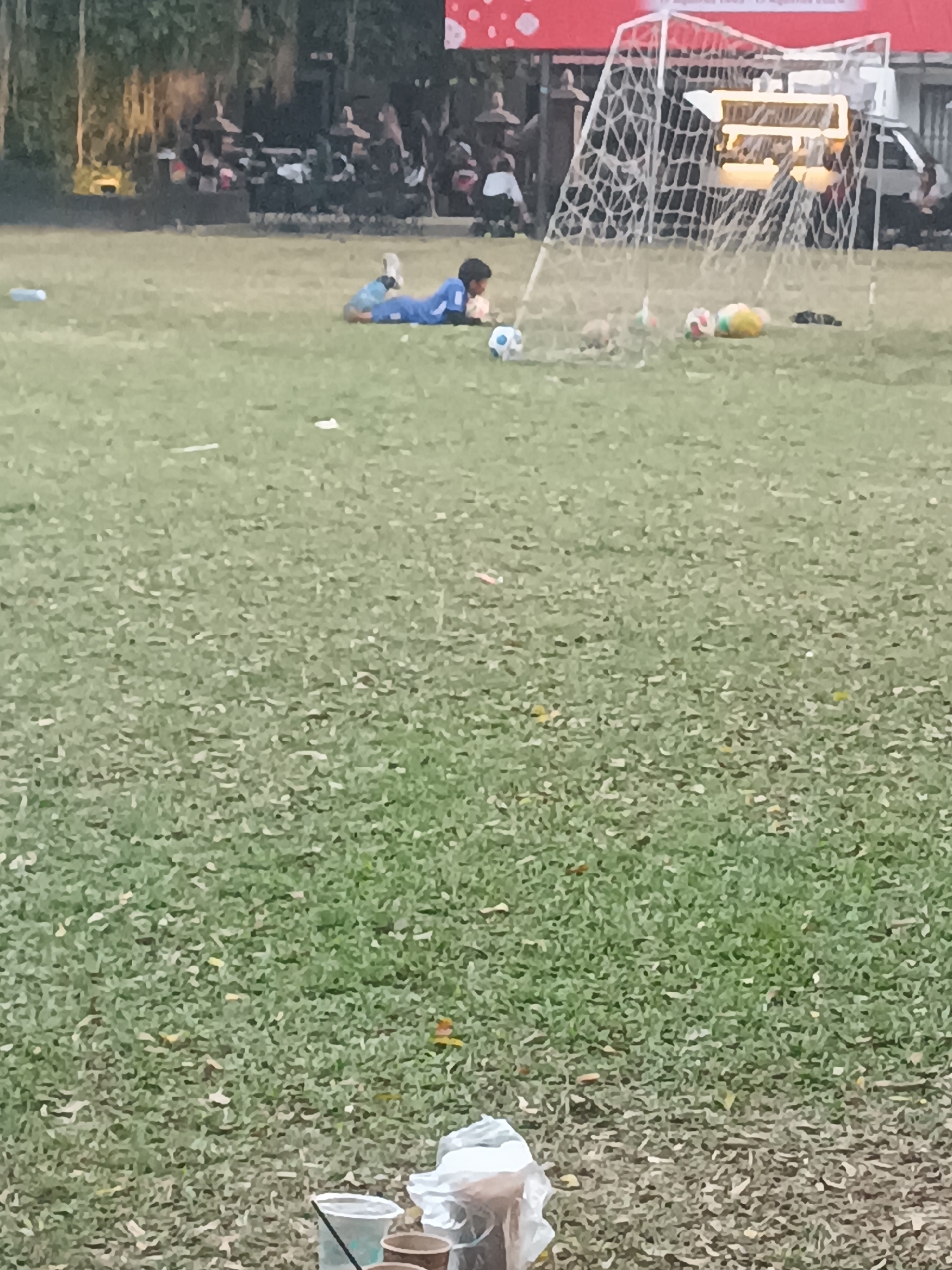 In the city of Sleman, Indonesia, a young person in a blue soccer uniform is lying on a grassy field in front of a soccer goal. Several soccer balls are scattered on the field near the goal. In the background, a crowd of people are gathered, and a vehicle is parked near the soccer goal. The scene appears to be a soccer practice or a casual game.