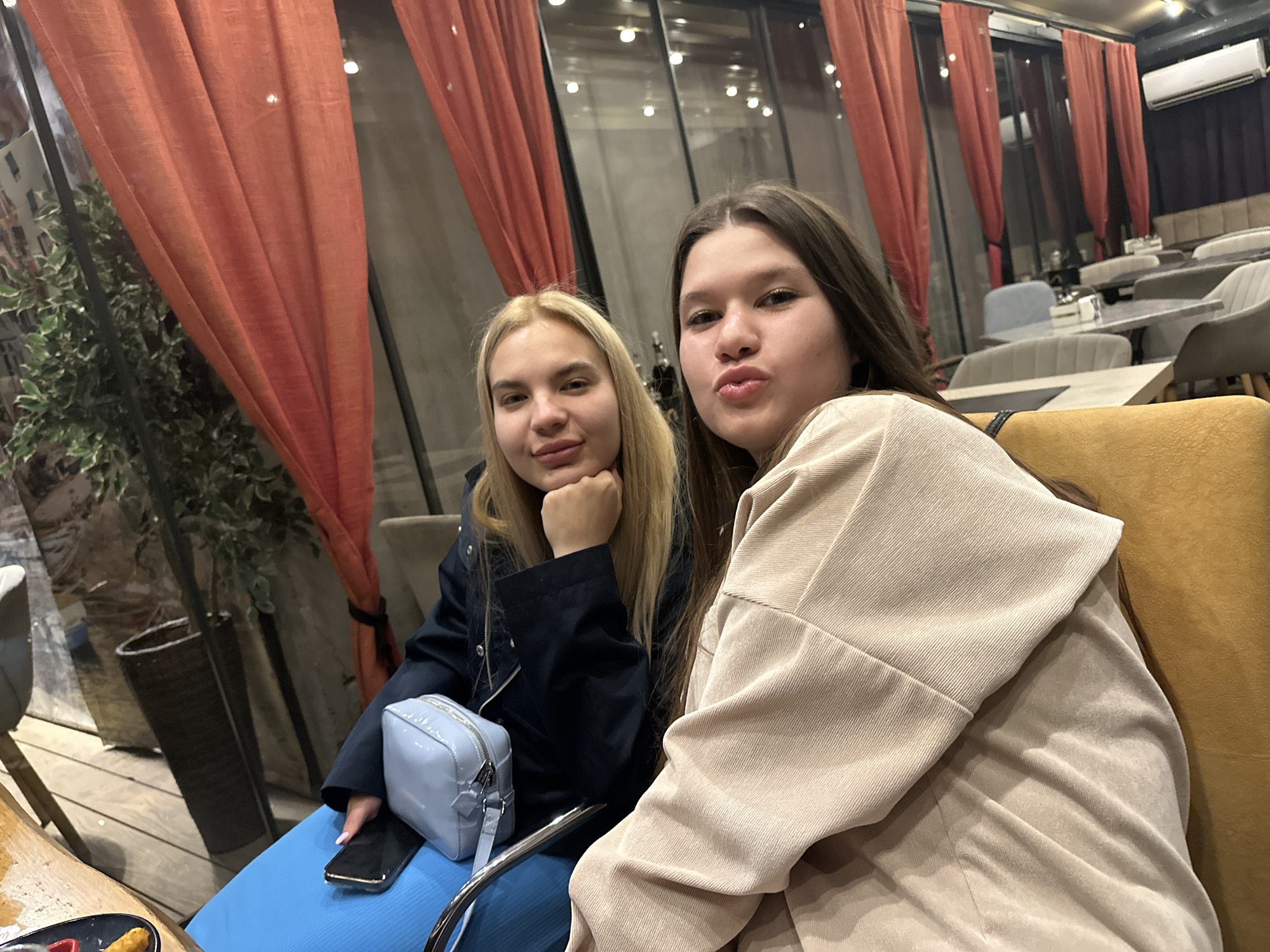 Two young women are posing for a photograph inside a restaurant. The woman on the left has blonde hair and is wearing a navy jacket, blue top, and has a small light blue purse on her lap. The woman on the right has brown hair and is wearing a beige corduroy jacket. The restaurant has orange curtains, large windows, and other patrons are seated at tables in the background. The location is possibly in Sofia, Bulgaria.