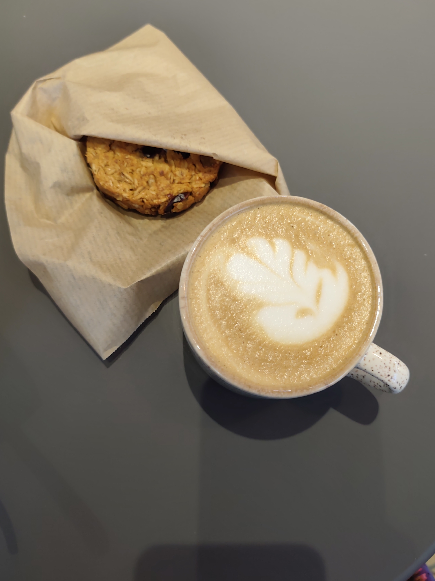 A round, oatmeal and raisin cookie is peeking out from a light brown paper bag, resting on a dark grey surface. Next to the cookie, a white mug with speckled patterns contains a latte with foam art, shaped like a leaf. The background is a plain, light grey wall. The scene appears to be indoors, on a tabletop.