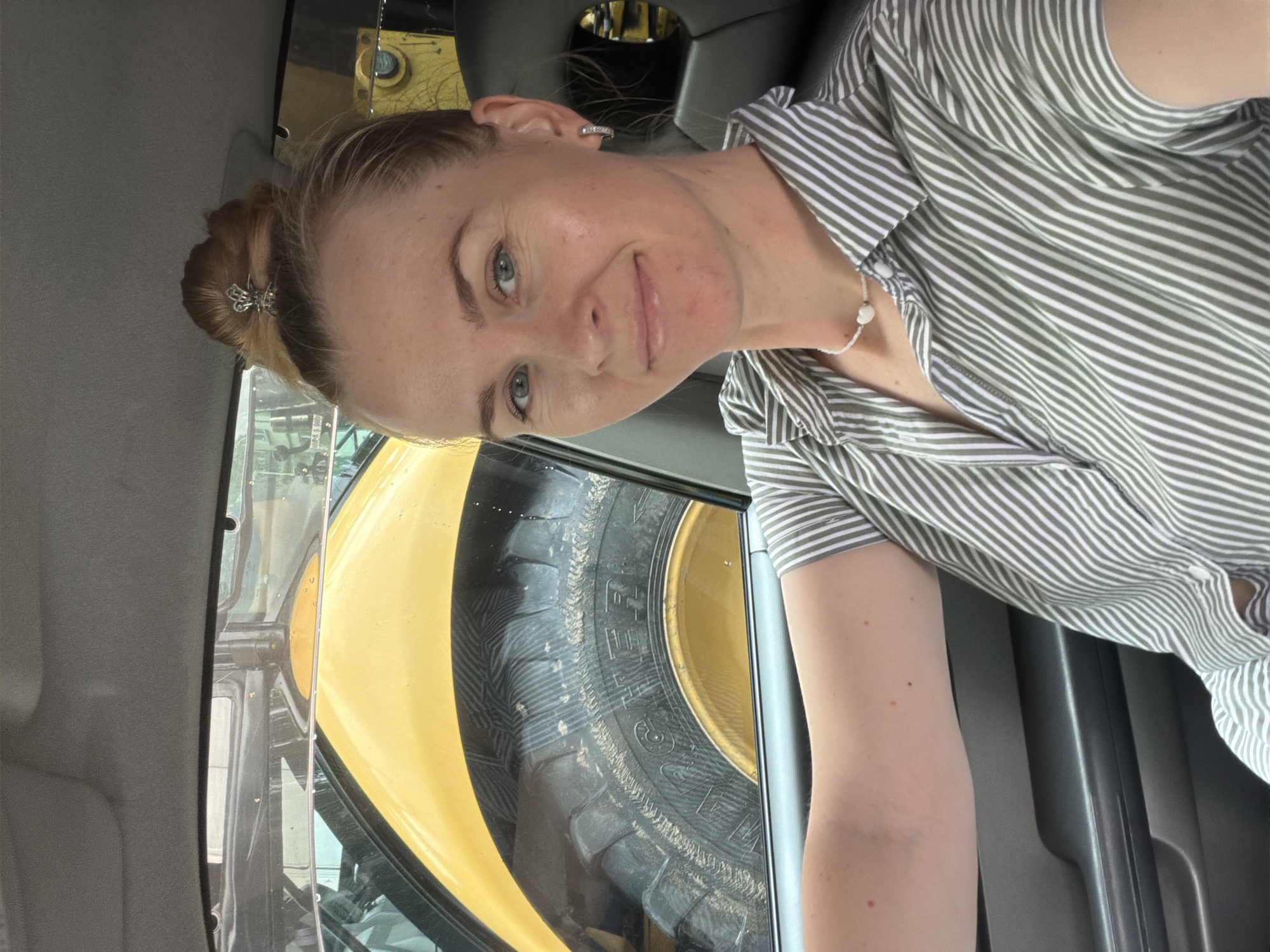 A blonde woman with her hair in a bun is sitting in the driver's seat of a vehicle. She is wearing a green and white striped shirt and a white necklace with a heart pendant. The woman is smiling and looking at the camera. Through the car window, a large yellow piece of heavy machinery with a large tire is visible. The scene appears to be outdoors, with natural light. The location is the city of Khabarovsk, Russia.