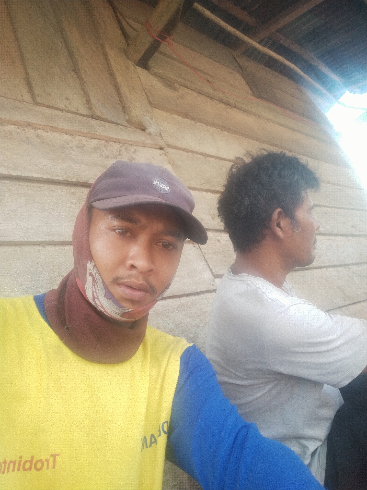 The image depicts two men in an outdoor, possibly rural, setting. The man in the foreground, wearing a yellow shirt and a brown cap, is looking directly at the camera. He has a mustache and is wearing a patterned cloth wrapped around his head and neck. The man in the background is wearing a gray t-shirt and is looking to his right. They are seated in front of a wooden wall. The overall impression is that of a casual or work-related break.
