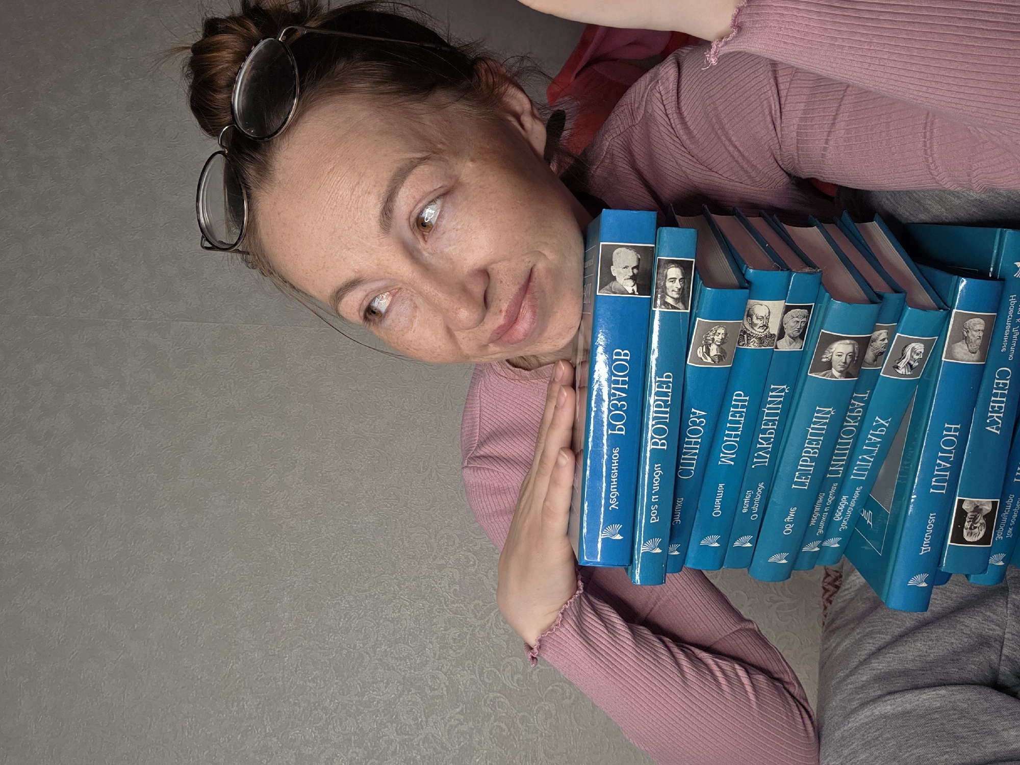 The image shows a woman with brown hair pulled into a bun, wearing glasses perched on top of her head and a long-sleeved pink shirt. She is holding a stack of blue books with portraits and Cyrillic text on the spines. The books feature titles such as "ВОЗУНОВ", "ВОИРЛЕБ", "СЕНЕКУ", and names like "МОНЕР", "ЛЕЙБВЕПΝΝ", and "НОТАПП". The text on the books seems to be in Russian. The background is a gray, textured wall. Based on the Cyrillic script on the books, it can be inferred that the location might be somewhere where Russian is spoken or published. As the books are in Russian and appear to be of literary or intellectual content, the location is most likely somewhere in Russia. However, without additional information from the image itself, it is impossible to determine if the location is in the city of Chaikovsky, Russia.