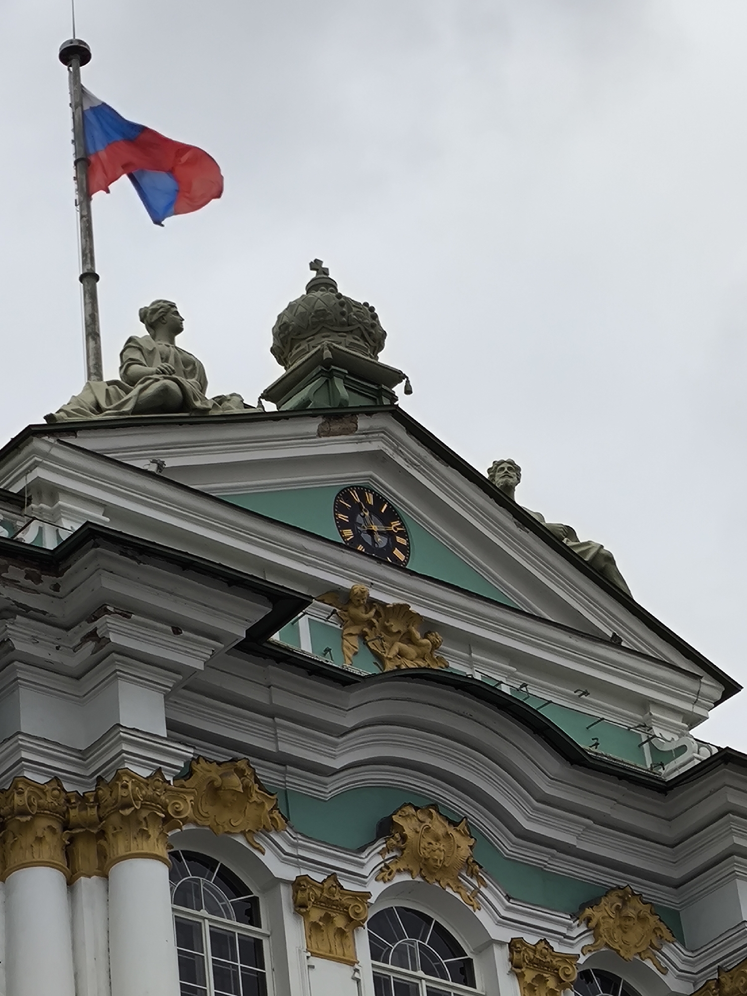 The image depicts the upper facade of the Winter Palace in Saint Petersburg, Russia, on an overcast day. A Russian flag, with red, blue, and white horizontal stripes, flies from a tall flagpole on the left. The flag is partially unfurled and appears to be waving.
The palace facade is ornate, featuring classical architectural elements. Two stone statues of seated figures adorn the roofline. On the left, a female figure is depicted with flowing drapery. On the right, a male figure with a beard is partially visible, looking upwards. Above the central triangular pediment, a large, ornate crown, likely a representation of imperial regalia, sits atop a decorative structure.
A large circular clock with Roman numerals is prominently featured on the pediment. Below the clock, a golden sculptural element depicts cherubic figures and ornate scrollwork. The building's facade is painted in a pale green and white color scheme, with decorative gold accents on the cornices, columns, and sculptural elements. The windows visible are arched and divided by mullions. The overall impression is one of grand, historical architecture under a cloudy sky.