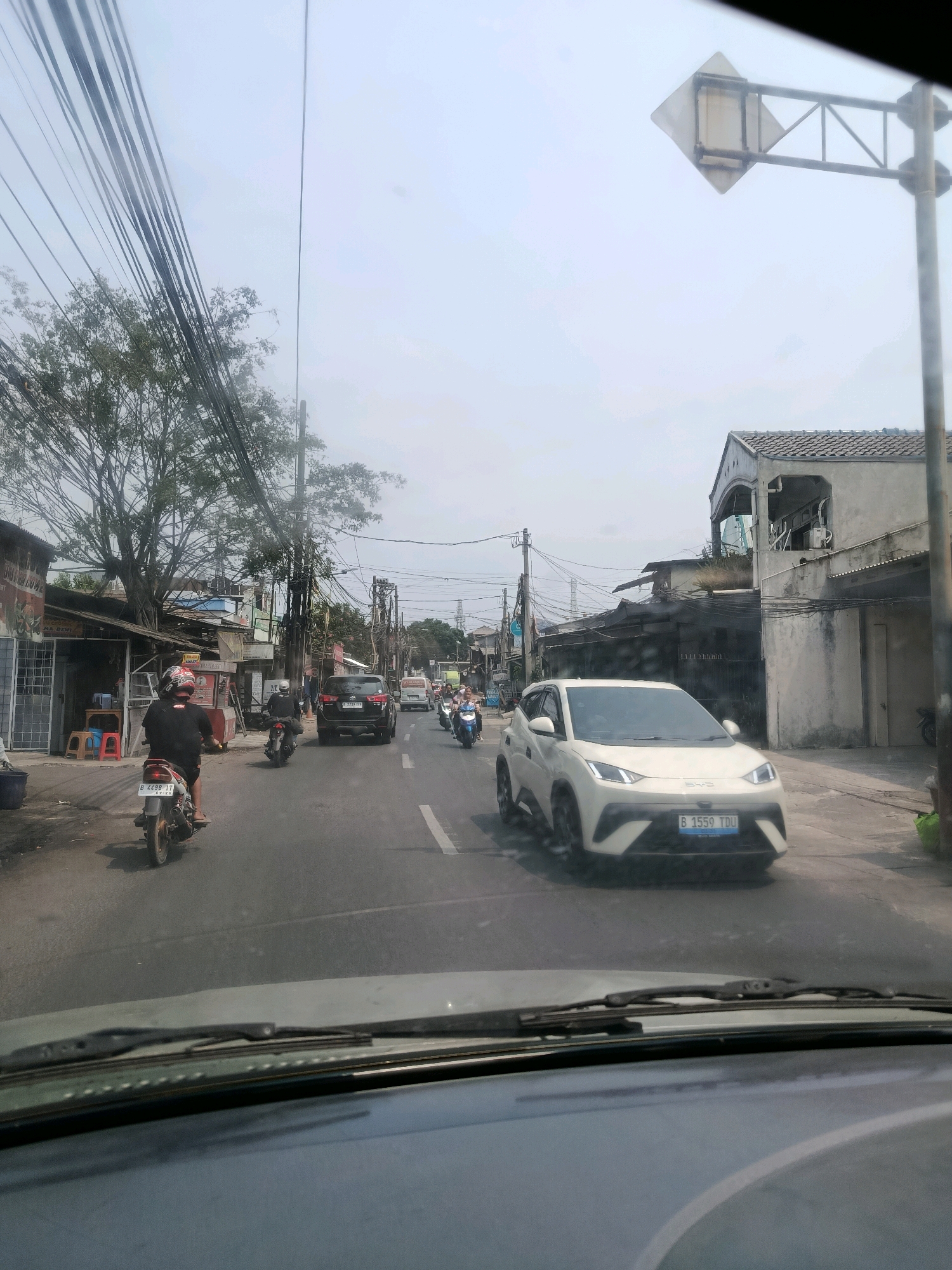 The image captures a street scene in East Jakarta, Indonesia, likely during daylight. The perspective is from inside a vehicle, looking forward through the windshield. The street is paved and appears to be a two-lane road with a painted white line dividing the lanes.
Several vehicles are present: a white SUV is prominent in the foreground on the right lane, with its license plate reading "B 1559 TDU". To the left of the SUV, a black SUV is in the left lane. Several motorcycles are also on the road. In the foreground left lane, a motorcycle with a rider wearing a black shirt and helmet is visible, its license plate displaying "B 4498 LT". Further down the road, more motorcycles are seen.
The environment features a mix of residential and commercial buildings lining the street. On the left side, there are shops with open fronts, some displaying signage, and a large tree with dense foliage overshadowing the street. Utility poles and a complex network of electrical wires are strung overhead, a common sight in urban Indonesian landscapes. On the right side, there are buildings with tiled roofs and balconies, typical of the architecture in the region. A diamond-shaped traffic sign is mounted on a pole on the right side of the road, facing away from the camera. The sky is overcast, giving the scene a diffused light. The overall impression is of a busy, typical urban street in a developing city.