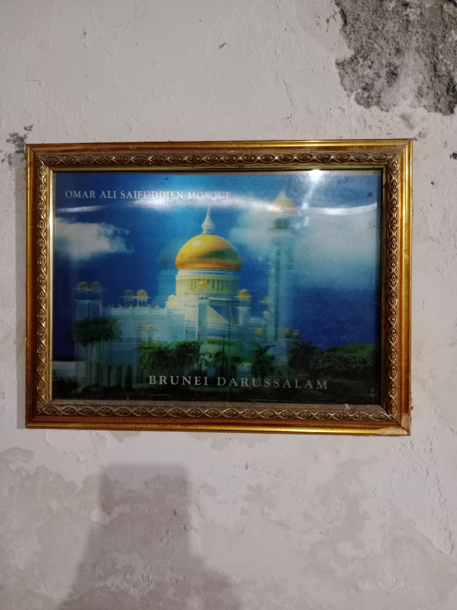 A framed photograph of the Omar Ali Saifuddien Mosque in Brunei Darussalam is displayed on a textured, off-white wall with visible patches of dampness and peeling paint. The photograph, enclosed in a golden, ornate frame, depicts the mosque with its prominent golden dome against a blue sky with white clouds. The mosque appears to be illuminated, casting a bright light. Trees and foliage are visible in the foreground of the photograph. The text "OMAR ALI SAIFUDDIEN MOSQUE" is printed across the top of the image, and "BRUNEI DARUSSALAM" is printed across the bottom. The wall's texture suggests it is concrete or plaster, and the damp areas indicate potential water damage. The lighting in the scene is ambient, coming from the top left.