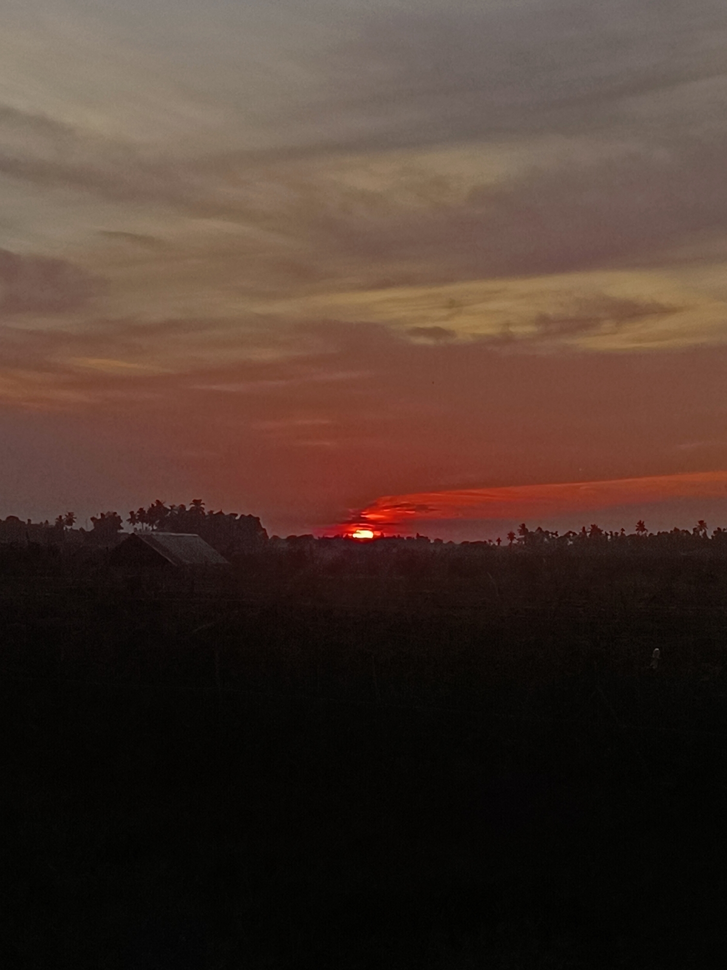 The image shows a sunset over a rural landscape in Meunasah Bie, Indonesia. The sky is filled with hues of orange, red, and gray, with clouds casting shadows. In the foreground, a dark silhouette of vegetation and a small structure are visible. The sun, a bright orange-red disk, is partially obscured by a low-lying cloud bank on the horizon. Palm trees and other foliage form a dark line against the vibrant sky. The overall scene depicts a serene and tranquil evening in a tropical setting.