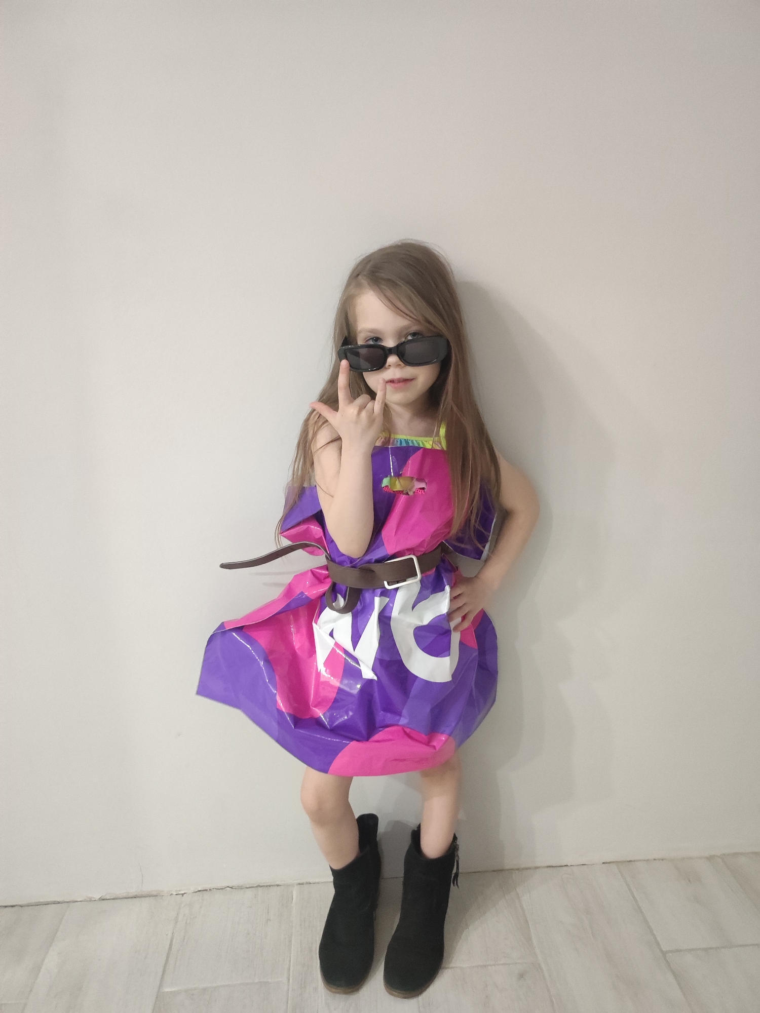 A young girl stands against a white wall, posing in what appears to be a homemade dress crafted from a Taco Bell paper shopping bag, cinched at the waist with a brown belt. She is wearing dark sunglasses and black ankle boots, striking a confident pose with one hand on her hip and making a rock-on gesture with the other hand. The environment is a simple room with light-colored wood-like flooring. The photo was taken indoors, and there's no clear indication of the location being in Votkinsk, Russia.