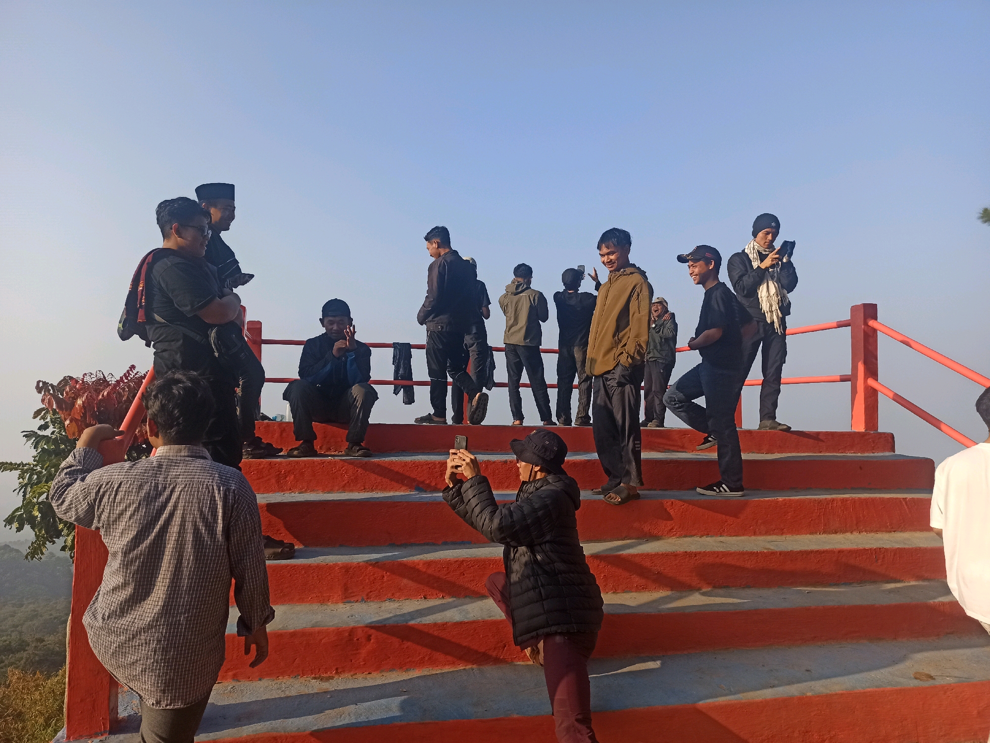 In Cileunyi, Indonesia, a group of people are gathered on bright orange stairs leading up a hill. The sky is a clear, pale blue, indicating a bright, sunny day. In the foreground, a person wearing a checkered shirt faces away from the camera, looking out at the landscape. To their right, another person kneels on the stairs, holding a smartphone and appearing to take a photograph. Further up the stairs, several individuals stand and observe the surroundings, with some holding phones and one man appearing to be taking a selfie. The stairs are bordered by a red railing. The environment suggests an outdoor scenic viewpoint.