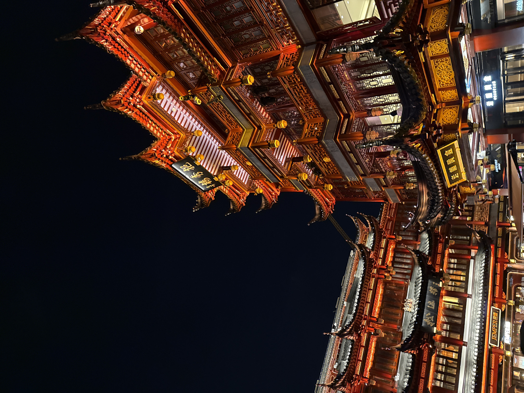 This image depicts the exterior of traditional Chinese architecture illuminated at night in Shanghai, China. The buildings are adorned with red and gold detailing, intricate carvings, and multiple tiers of ornate roofs. The dominant structure on the right is a multi-story building with detailed wooden latticework and decorative lanterns hanging from the eaves. A sign with Chinese characters is visible on the upper part of this building. To the left, another building with similar architectural elements is visible, also lit with red lights. A sign that reads "JEMPER" is present on the lower level of the left building. The sky is a deep, dark blue, indicating nighttime. The scene appears to be a bustling commercial or cultural district, possibly a market or a historical area. Several people can be seen on balconies and walkways of the buildings, engaged in everyday activities, though their features are not distinct due to the distance and lighting. The overall impression is of a vibrant and beautifully illuminated historical or culturally inspired area within Shanghai.