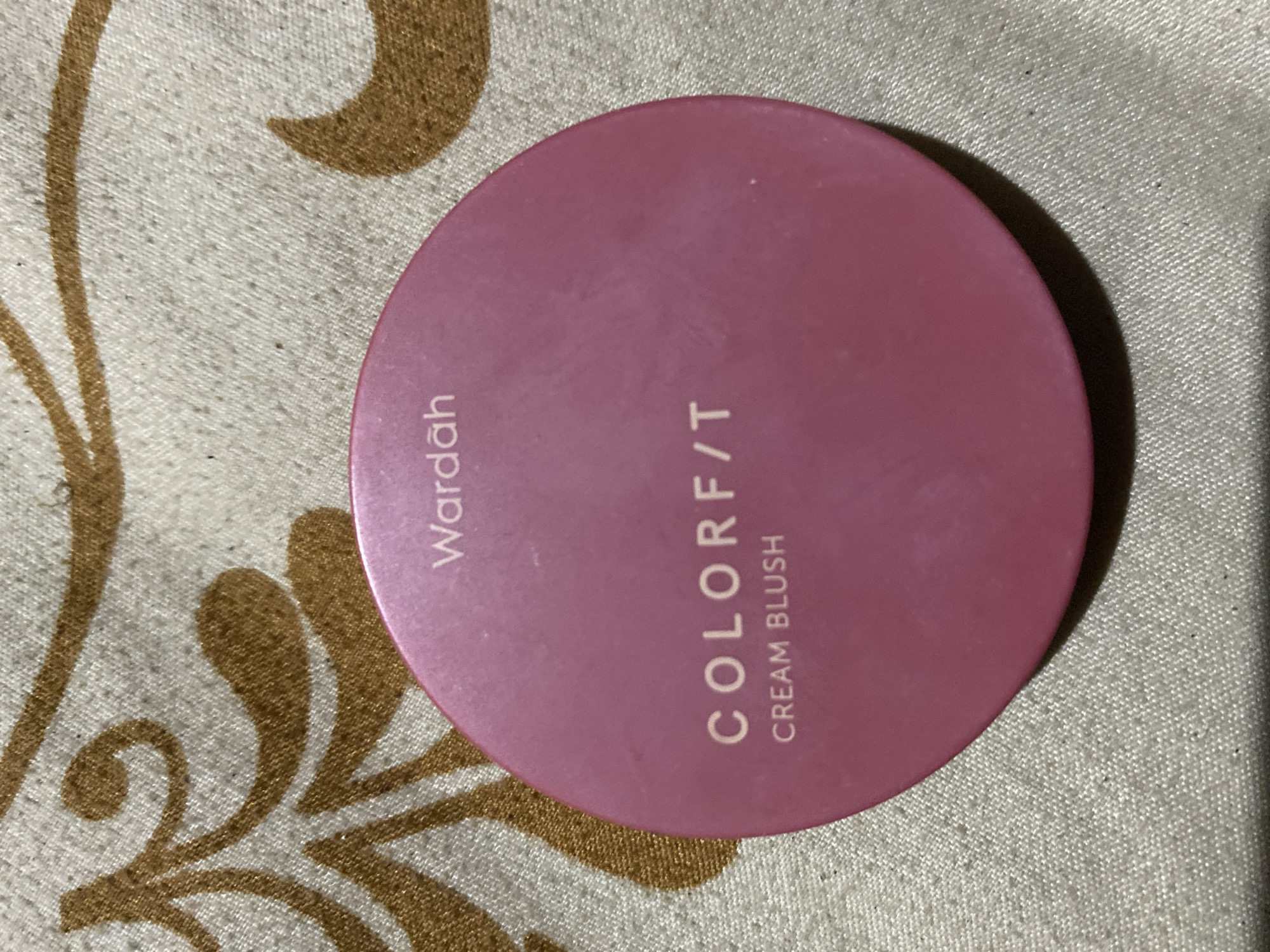 The image shows a pink circular makeup compact, specifically a Wardah Colorf/t Cream Blush, resting on a textured fabric surface with a brown decorative pattern. The product packaging is prominently displayed, with the brand name "Wardah" and the product name "COLORF/T CREAM BLUSH" visible in white lettering. The fabric appears to be a tablecloth or cushion cover. There are no people or any indications of a specific location within Ta', Indonesia. The lighting suggests an indoor setting.