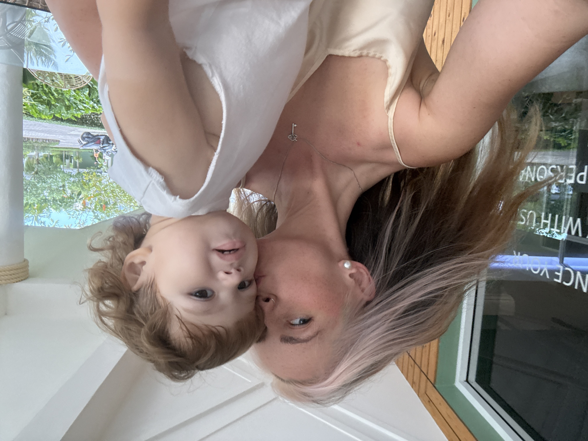 A woman with light brown hair streaked with pink kisses a young child on the cheek. The woman is wearing a beige satin camisole and a silver necklace with a key pendant. The child is wearing a white sleeveless top. They are outdoors, possibly on a patio or balcony, with a view of greenery, a road, and buildings in the background. The environment appears tropical, and the presence of a motorcycle suggests a location in Thailand. Specifically, the signage visible through a window in the background, which reads "NCE YOUR" and "WITH US" and "PERSONA", suggests a business establishment in Choeng Thale, Thailand.