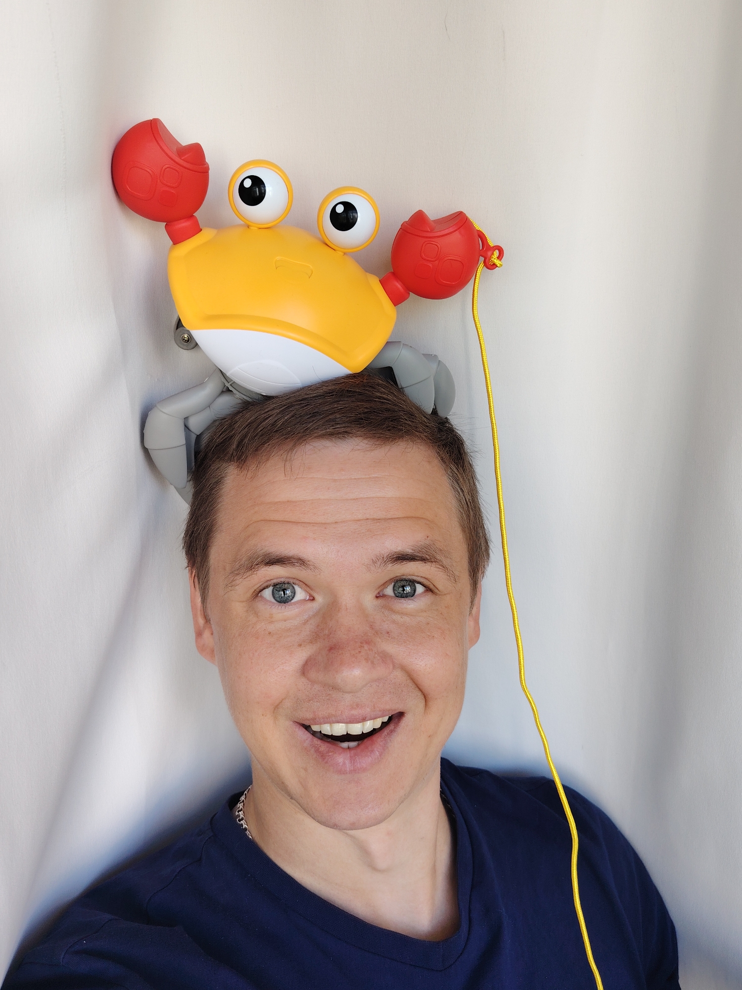 A smiling man with blue eyes and short brown hair poses for a selfie indoors with a yellow and red toy crab perched on his head. The man is wearing a navy blue t-shirt and a silver chain. The crab toy has googly eyes, large red claws, and a yellow pull string. The background is a plain white wall with subtle vertical creases, suggesting a curtain or draped fabric. The setting is likely a domestic environment, potentially in Chaikovsky, Russia.