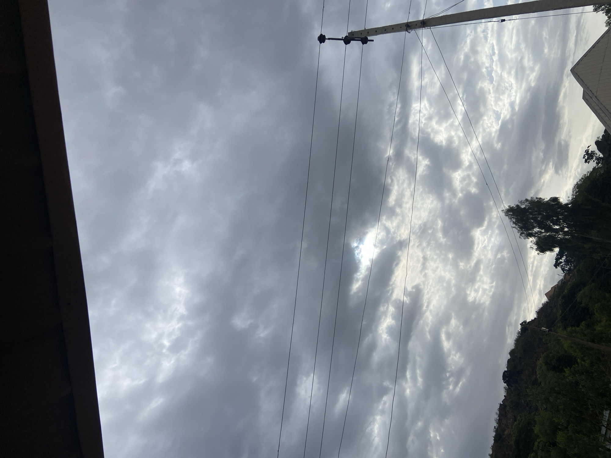 The image shows a cloudy sky with a utility pole and power lines running across the frame. Part of a building's eaves is visible on the left. A hillside covered in trees is on the right side of the image. There is a small glimpse of sunlight peeking through the clouds. The image was taken from Jos, Nigeria. There are no people visible.