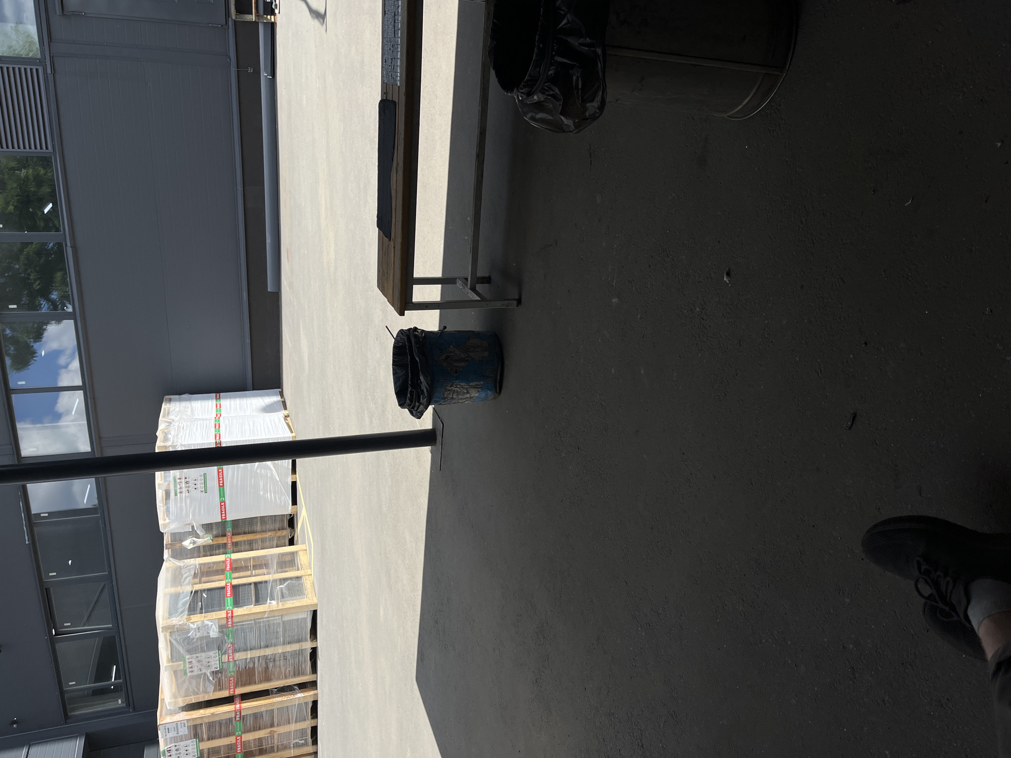 The image depicts an outdoor industrial or warehouse area with a concrete floor and a dark grey building in the background. Several large, shrink-wrapped pallets are stacked on the left side of the frame, some with visible "FRAGILE" tape. A wooden bench with a metal frame is positioned in the middle of the scene, with a black mat on its surface. Two trash cans are present: one blue, weathered bin is to the left of the bench, and a larger, dark metal bin with a black liner is to the right. The ground is mostly concrete, with some areas in shadow cast by an unseen overhead structure or the building. The lighting suggests it is daytime. There are no people clearly visible, though a person's legs and feet, wearing dark athletic shoes, are present in the lower left corner of the frame, indicating someone is seated or resting. The scene appears to be an outdoor staging or storage area. There is no identifiable information in the image that would place it specifically in Kyiv, Ukraine.
