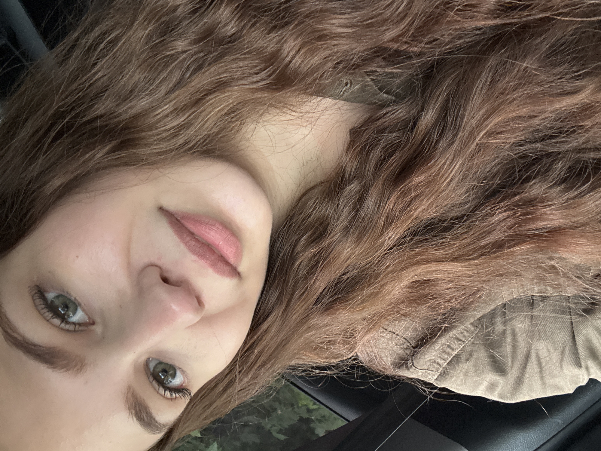 This is a close-up, sideways selfie of a young person with long, wavy, light brown hair. The person's face is partially visible, with their head tilted. They have fair skin, light green eyes with visible eyelashes, and light brown eyebrows. Their lips are painted with a matte, rosy-brown lipstick, and they appear to be wearing a tan-colored jacket or shirt. The background is blurred, showing hints of dark green foliage, suggesting an outdoor setting, possibly in a car given the dark interior visible at the bottom right. The lighting is soft, indicating it might be daytime, perhaps overcast or late afternoon. The overall mood is calm and relaxed. No text is visible in the image. The location context provided is Chaikovsky, Russia.