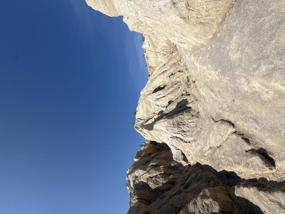 The image depicts a deeply eroded geological formation under a clear blue sky in the city of Druzhba, Ukraine. The environment is an outdoor, arid or semi-arid landscape dominated by extensive erosion. The sky is a cloudless, vibrant blue, occupying the upper third of the frame. The terrain consists of light beige to off-white sedimentary material, likely clay or sand, forming steep, jagged ridges, peaks, and pronounced valleys. Distinct vertical striations and gullies are visible on the eroded surfaces. A relatively flat, light-colored path or channel meanders through the foreground and into the midground. Sparse, small clusters of green vegetation are present on the upper edges and crests of some formations. Indistinct etched or carved markings, possibly alphanumeric characters, are visible on the surface of the large formation in the right midground. The scene is illuminated by direct, bright sunlight, casting sharp shadows that highlight the contours of the landforms. No people, specific actions, or interactions are present in the frame.