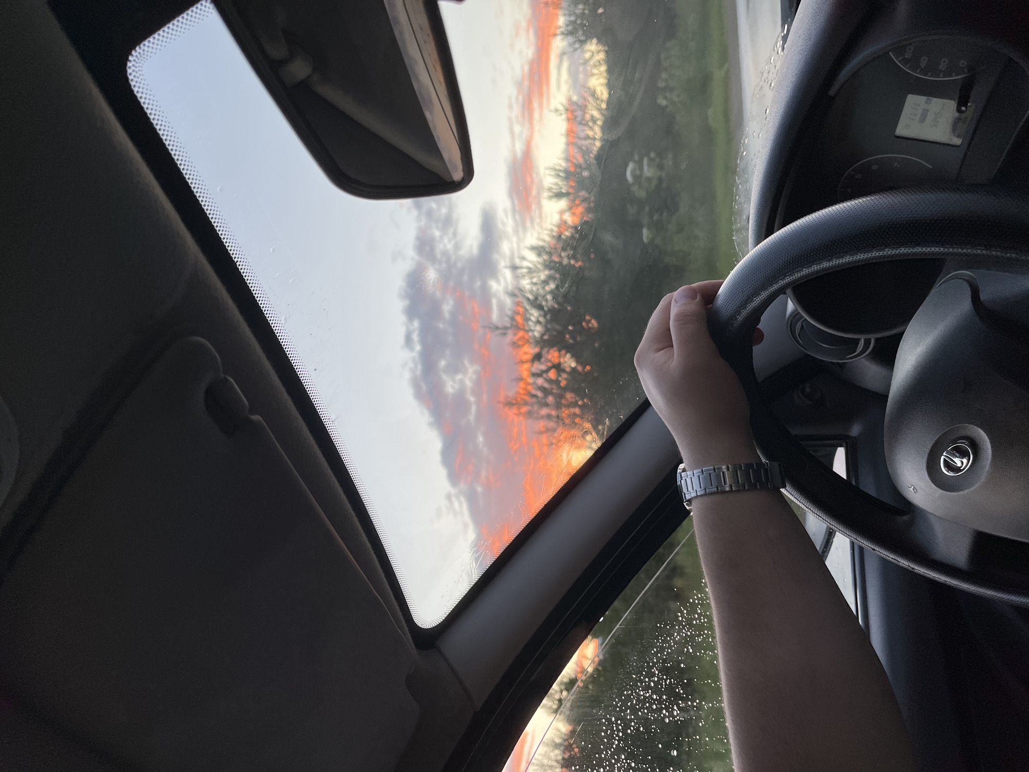The media file shows the interior of a car during sunset. The focus is on a person's arm and hand holding the steering wheel, with a wristwatch visible on their wrist. The car's dashboard and a portion of the steering wheel are in the foreground. Through the windshield and sunroof, a vibrant sunset with orange and pink clouds is visible, set against a backdrop of trees. The sky is bright, indicating either sunrise or sunset. The weather appears to be clear, though there are some water droplets on the windshield, possibly from recent rain or condensation. The overall mood is peaceful and serene, capturing the beauty of the natural light. No visible text is present in the image. The location is identified as Prikamsky, Russia, suggesting a driving scene in that region.