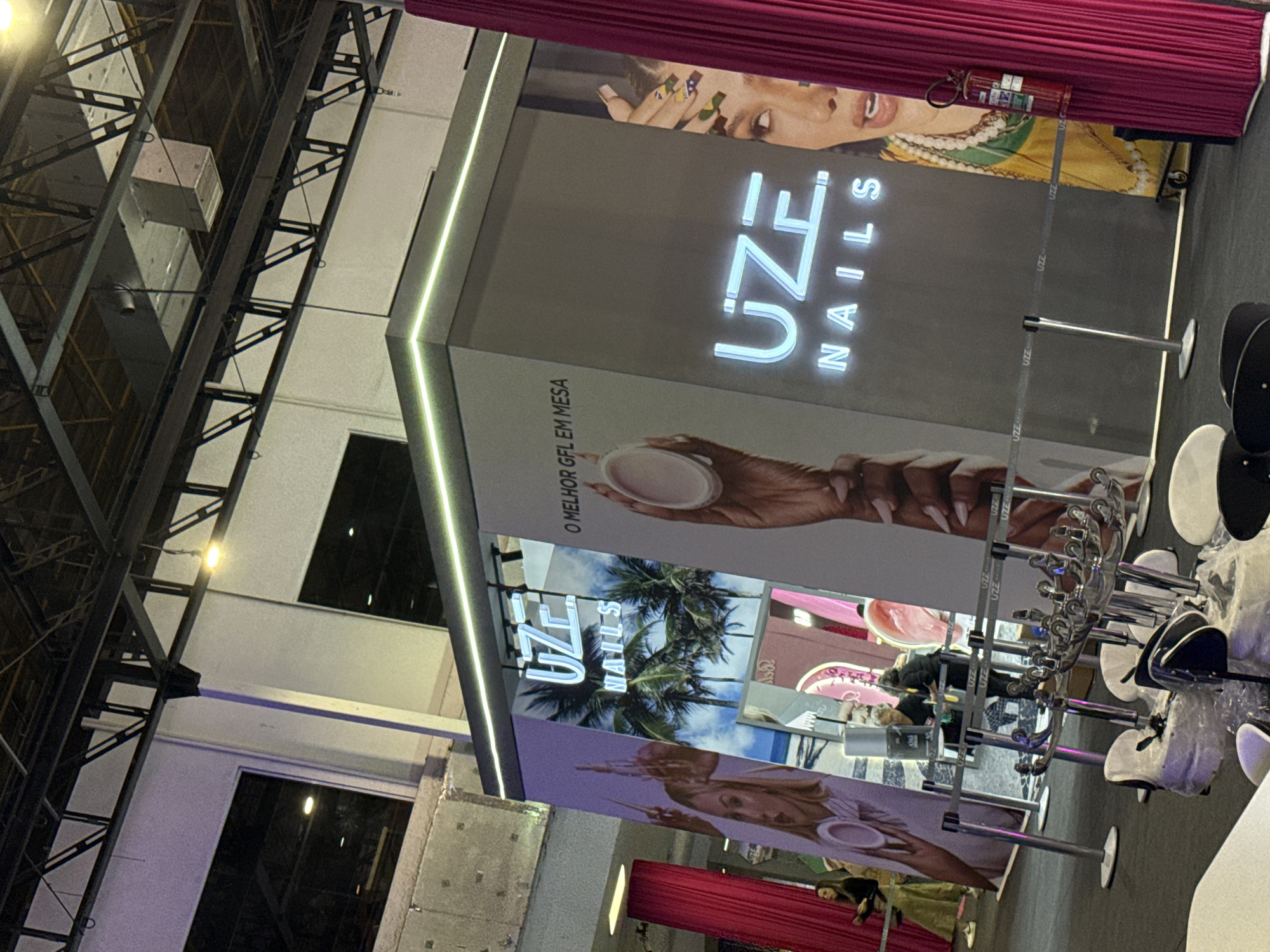 This image depicts a commercial booth for "UZE Nails," likely at a trade show or similar event. The booth features large vertical displays adorned with photos and the brand name, written in a luminous sans-serif font. The booth is lit from within and topped with strips of LED lights. The display materials include nail care products, a diverse collection of manicures, and palm trees. Metal stanchions and swivel chairs are in the immediate foreground. In the background, people can be seen within the booth area. Architectural elements, including metal framing, are present in the upper left portion of the frame. Based on the image content (UZE Nails is a Brazilian brand), the location is most likely in the city of São Paulo, Brazil.