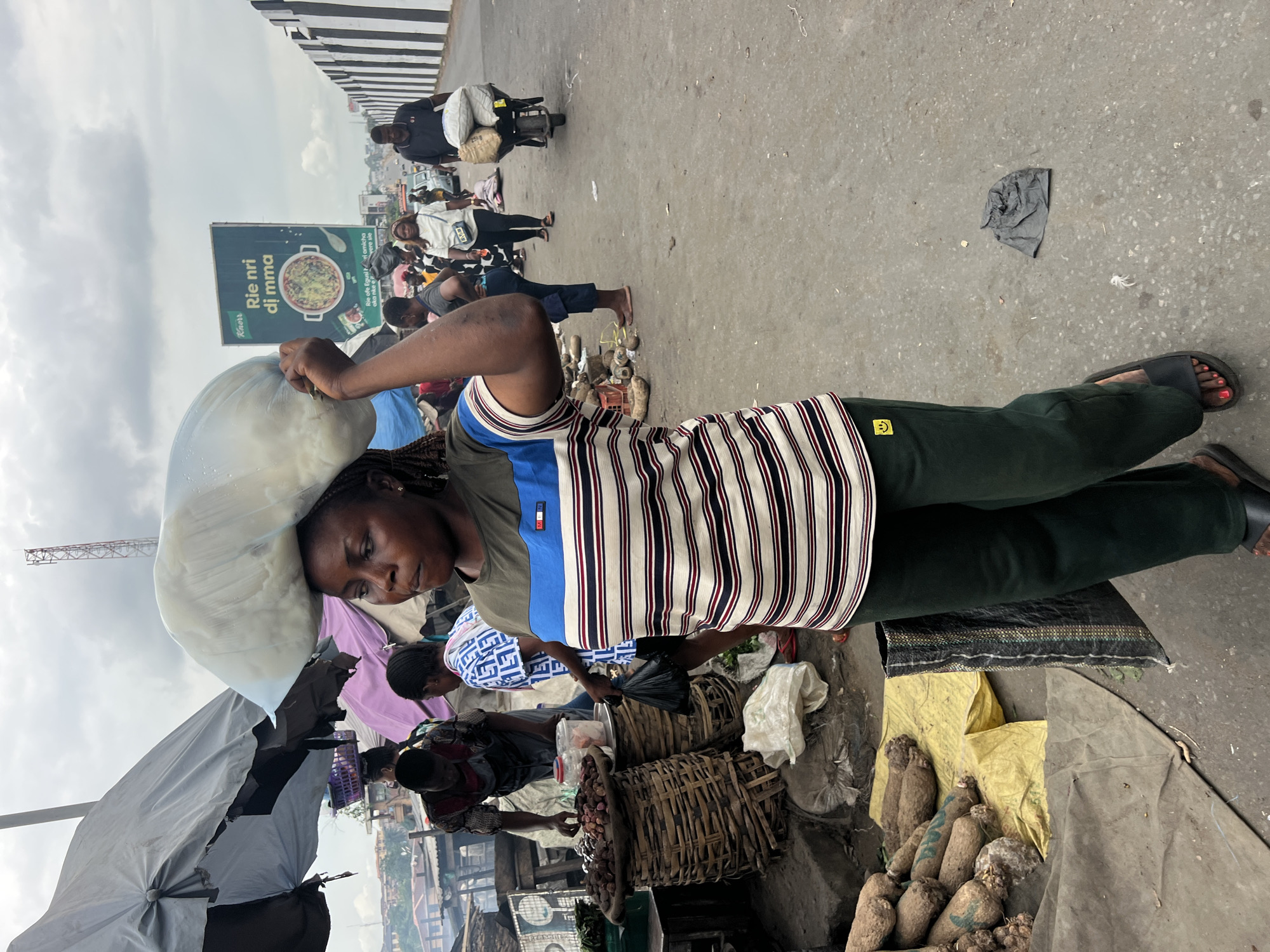 This medium shot captures a bustling outdoor market scene, likely Nkpor, Nigeria, under an overcast sky. In the foreground, a woman carries a large, light-colored sack on her head, balancing it with her right hand. She wears a striped short-sleeved top with blue, white, and dark red stripes, dark green pants featuring a yellow smiley face patch on the left thigh, and black sandals with red nail polish visible on her toes. She looks slightly to her left, appearing focused.
Behind her, the market is active. Several other individuals are present; another woman in a patterned blue and white top attends to goods in woven baskets. Heaps of yams or similar root vegetables are displayed on yellow sacks and grey tarps on the ground. An open grey umbrella provides shade for a stall on the left. In the mid-ground, a man rides a motorcycle, laden with packages, moving along the paved road. Further back, more people are visible, and urban buildings, including a communication mast, dot the distant skyline.
A prominent billboard in the middle distance features an image of a bowl of food. The brand "Knorr" is visible, and the main text reads "Rie nri dj mma," with smaller, partially obscured text below it, likely in the Igbo language. The scene conveys the busy, functional atmosphere of a typical West African street market. The sky is heavily clouded, suggesting an overcast day.