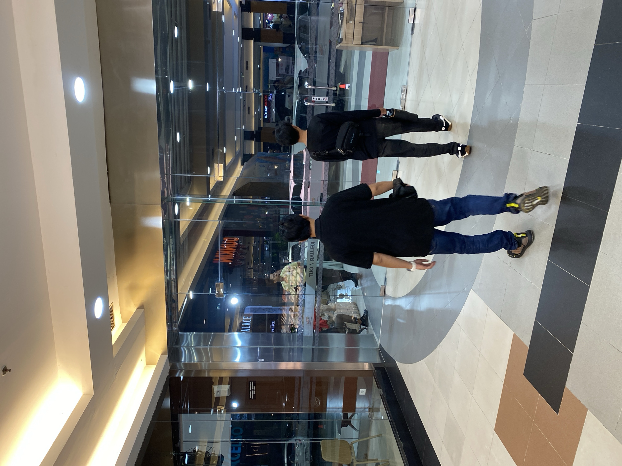 In Banjarmasin, Indonesia, two individuals, both appearing to be young men, are walking away from the camera in an indoor commercial setting. The man in the foreground is wearing a black t-shirt and blue jeans, with a black bag over his shoulder. He has on casual shoes with a yellow accent on the heel. The man behind him is wearing a black long-sleeved shirt and dark pants, with a black crossbody bag. He is also wearing casual shoes. They are walking on a tiled floor with a patterned design.
The environment appears to be a shopping mall or a large retail space. To their left, there is a glass storefront with reflections showing various cars, suggesting a parking area or a section of the mall with vehicle displays. The storefront itself has signage that is partially visible, including the word "PALACE". Inside the storefront, a person in a yellow and white checkered shirt and dark pants can be seen standing behind a counter, facing away from the camera. Another person is seated in the storefront.
The lighting in the space is bright, with recessed lights and illuminated ceiling panels. The overall impression is a well-lit, modern commercial interior. The scene is devoid of any overt interactions between the two individuals and their surroundings, other than their movement through the space.