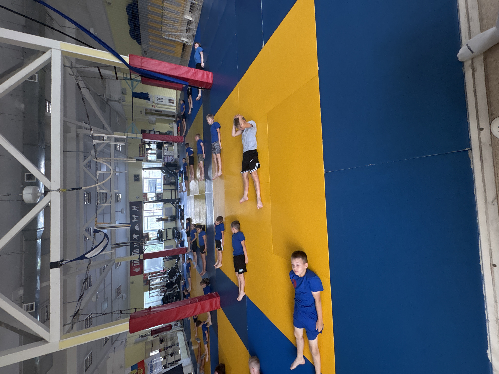 The image depicts a large, brightly lit gymnasium with a high ceiling and exposed structural beams. The floor is covered in a blue and yellow matting system. Numerous individuals, appearing to be children or young teenagers, are lying down on their backs on the mats, some with their arms at their sides and others with their hands behind their heads. They are dressed in athletic wear, primarily blue and black. In the background, there are gym equipment, weight machines, and banners or posters on the walls, one of which appears to have the Russian flag. There are also some circus-like apparatuses suspended from the ceiling, such as ropes and a blue fabric. The overall impression is of a group engaged in a physical training or stretching session. The presence of Cyrillic text on some of the posters suggests the location is in Russia. Given the details, this scene is likely taking place in a sports facility within the city of Chaikovsky, Russia.