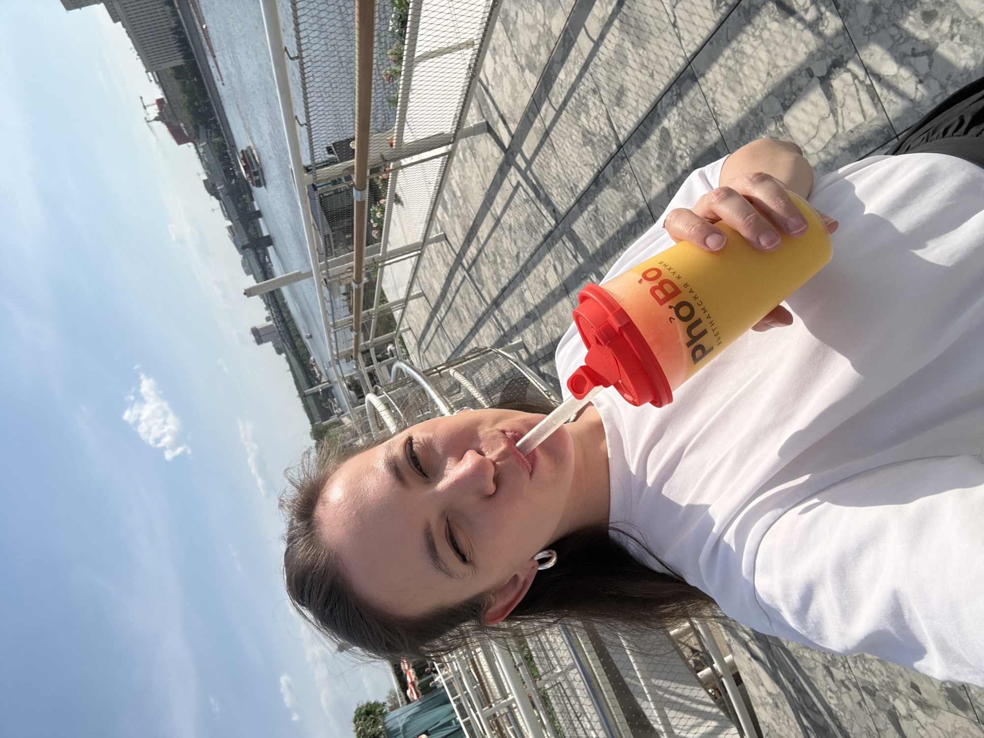 A person with long dark hair, wearing a white t-shirt, is drinking from a yellow and red cup through a straw. The cup has the inscription "Pho Bo" in Cyrillic alphabet. The person is standing on a paved surface next to a metal railing, with a river and buildings visible in the background. The location is Moscow, Russia.