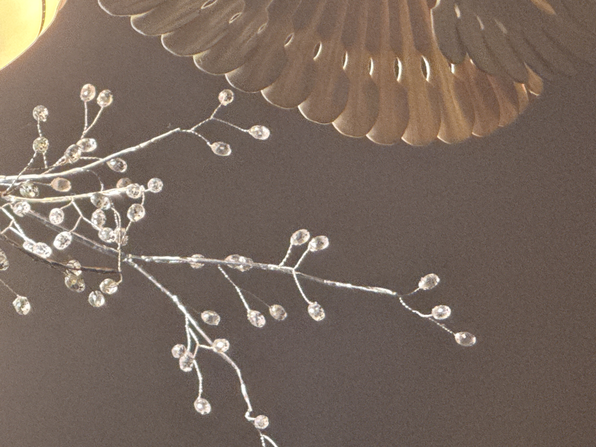 The image shows a close-up of a decorative object, likely a chandelier or a piece of jewelry, against a dark background. The object features delicate silver branches adorned with numerous clear, bead-like embellishments that resemble small crystals or pearls. These beads are attached to the branches by thin silver wires, creating a sparkling, almost ethereal effect. In the upper right portion of the image, a blurred element suggests a larger structure, possibly the source of light or a part of the main decorative piece, with a scalloped, layered design and a warm, golden hue. The lighting appears to be artificial, highlighting the reflective qualities of the beads and creating subtle shadows. There is no discernible text, activity, people, time of day, weather, or specific location cues evident in the image itself.