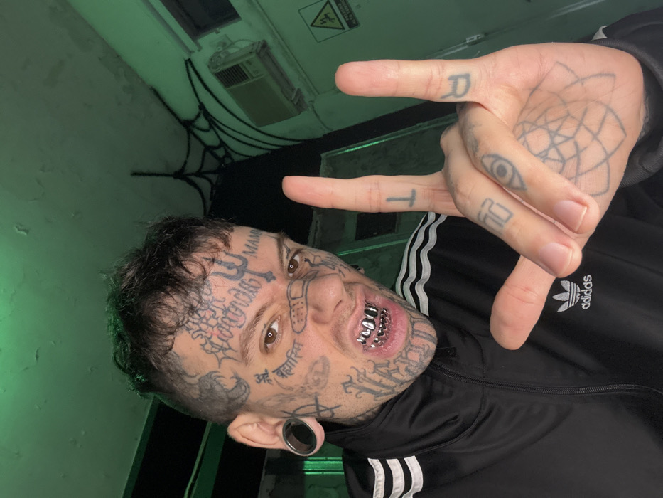 A heavily tattooed male subject, visible from the chest up, is illuminated by green lighting in an indoor setting. He has short, dark hair, large black ear gauges, and silver-colored grillz on his upper and lower teeth, bared in a grimace directed at the camera. His face is covered with numerous tattoos, including script on his forehead and cheeks, a trident on his forehead, an anarchy symbol, and a band-aid design across his nose bridge. He wears a black Adidas track jacket with white stripes on the sleeves and a white trefoil logo on the left chest. His right hand is raised, forming a "rock on" gesture, displaying further tattoos on his fingers (an inverted 'T' on the index, 'R' on the pinky) and a geometric pattern on his palm. The background consists of a light green-painted wall with visible electrical wiring and an old wall-mounted air conditioning unit. A dark, spiderweb-like structure is present on the upper wall/ceiling. A yellow triangular warning sign with a black lightning bolt symbol, indicative of high voltage, is partially visible on the right wall. The specific location within Buenos Aires, Argentina, is not discernible from the image.