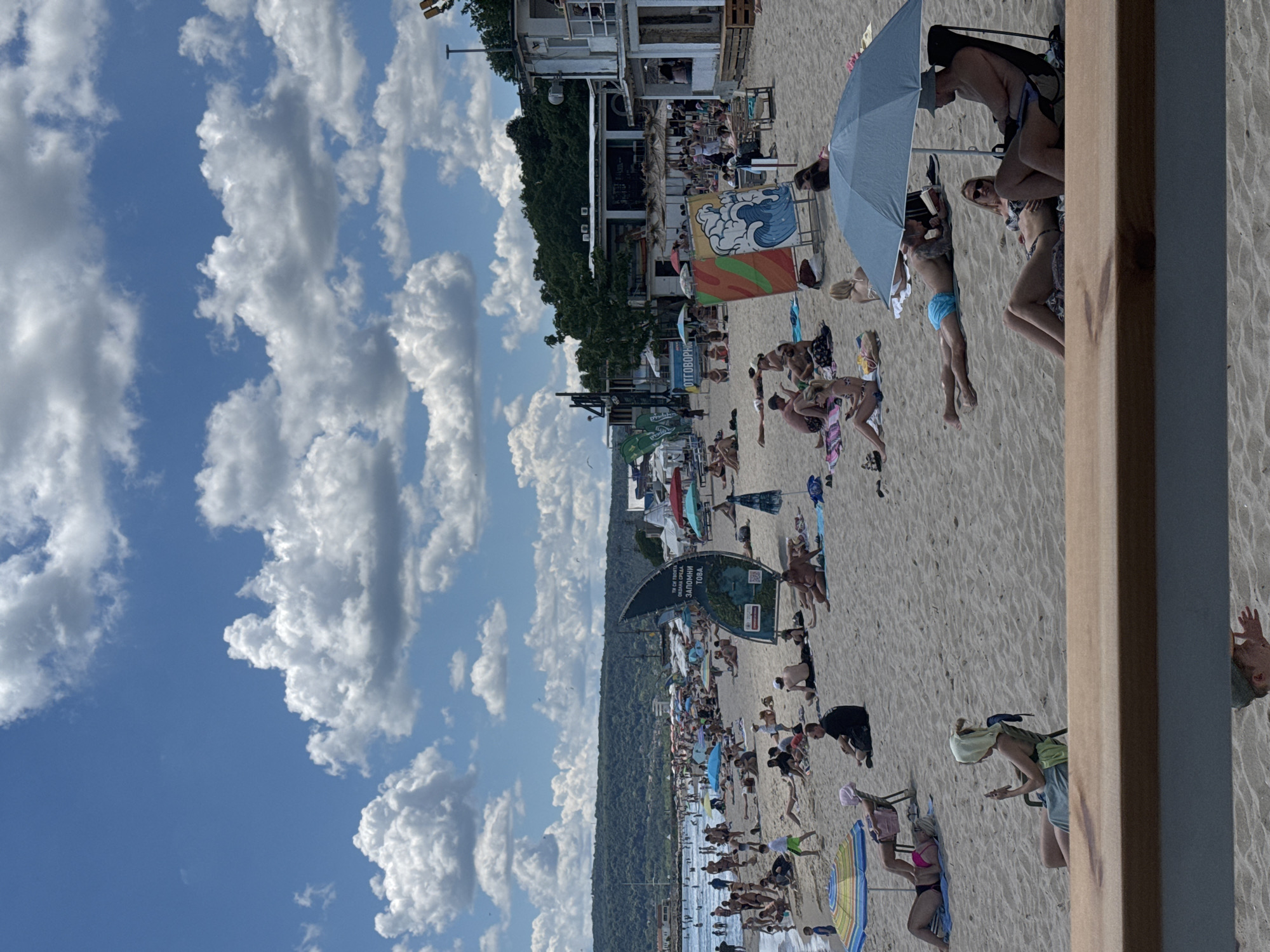 The image depicts a crowded beach scene on a sunny day, likely in Varna, Bulgaria, given the presence of Cyrillic text on a sign. The beach is sandy, with numerous people sunbathing, sitting on towels, and under umbrellas. Several beach umbrellas are visible, providing shade for the beachgoers. In the background, there are buildings, trees, and a hill. The sky is blue with scattered white clouds. Several people are in swimwear, enjoying the warm weather. Some are lying down, while others are sitting in beach chairs. A large, sail-like structure with text and images is positioned on the sand. The overall atmosphere suggests a typical summer day at a popular beach resort.
