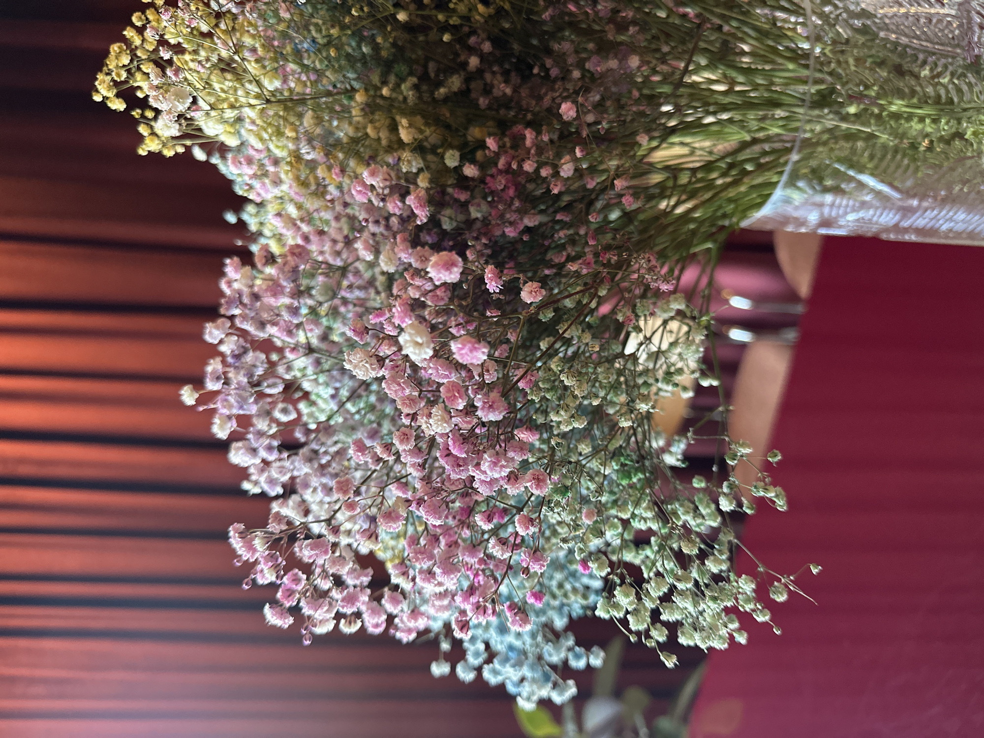 A close-up shot focuses on a bouquet of baby's breath flowers in various pastel colors, including pink, light blue, white, and pale yellow. The flowers are arranged in a clear glass vase, which is partially visible at the bottom right of the frame. Behind the flowers, a dark red curtain with vertical pleats serves as the backdrop. The lighting appears to be soft and natural, casting subtle shadows on the petals. The overall scene is serene and decorative, suggesting an indoor setting like a restaurant or home.