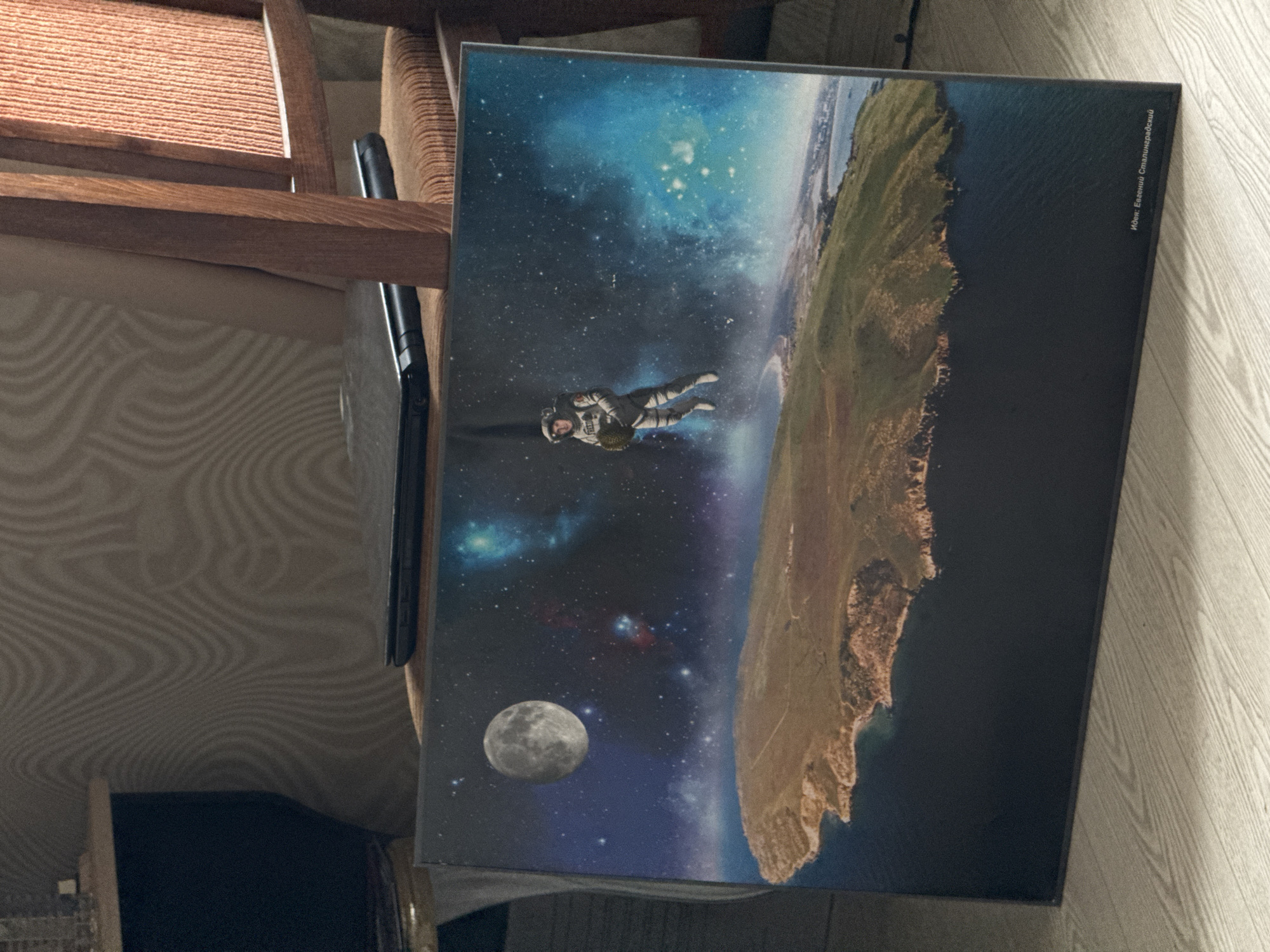 The image shows a poster or canvas print leaning against a light-colored wall. The print depicts an astronaut floating in space, with a view of a terrestrial landscape extending into the cosmos. The moon is visible in the lower left corner of the print. The upper right corner includes text: "Иван Евгеньевич Сталинградский". A black laptop is resting behind the print and on the chair, and part of a wooden chair is visible on the left side. The background includes a patterned wallpaper and part of a dark object, likely a piece of furniture. There is no visual indication of a location in the city of Ново-Никольское, Russia.