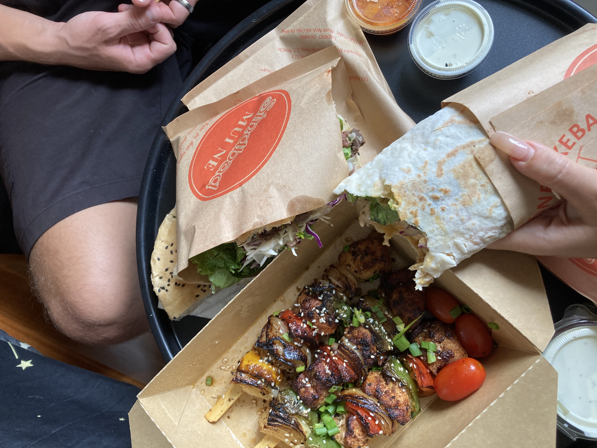 The image presents an overhead view of a meal featuring items from a restaurant named "Sindbad Mui Ne," as evidenced by the logo on a brown paper bag. The location is likely Mũi Né, Vietnam, given the restaurant's name. The meal includes kebabs on skewers garnished with sesame seeds and green onions, several red cherry tomatoes in the cardboard box. A person with light skin tone is holding a partially eaten wrap or kebab with their right hand. The wrap or kebab has a light-colored tortilla and some fillings with lettuce and purple cabbage. A man wearing a ring is on the left side of the image. A black tray containing multiple food items including containers with different condiments, and paper bags are visible.