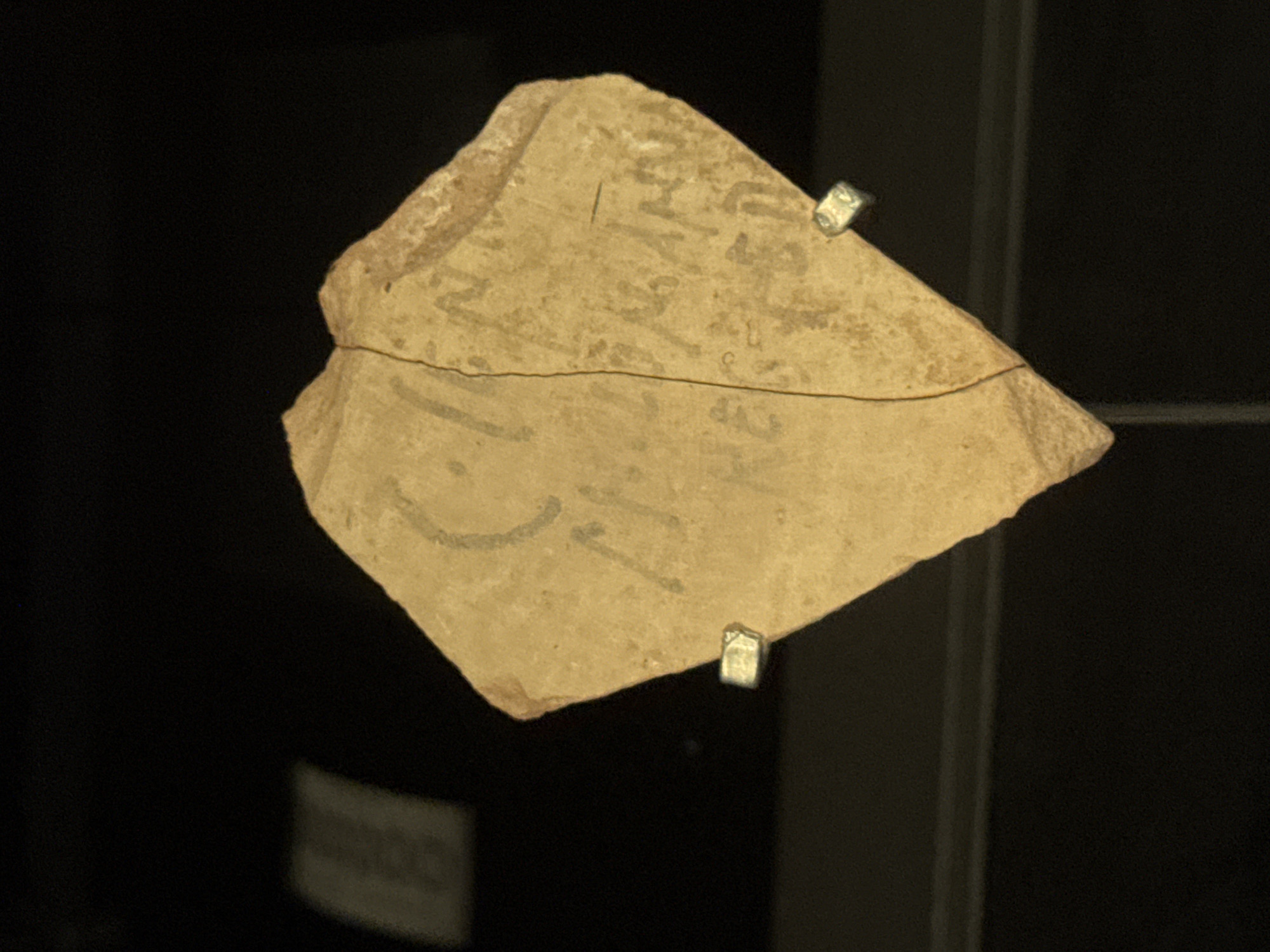 The image shows a shard of pottery with faint inscriptions, displayed in a museum setting. The pottery shard is light brown and has a cracked surface. Faint, dark markings resembling script are visible on the shard. Two small metal fixtures are attached to the shard, likely for display. The background is dark, with a blurred light-colored rectangular object visible to the left of the shard, suggesting it's on display in a case. The location is identified as Tamar Regional Council, Israel, suggesting the artifact is an archaeological find from that region.