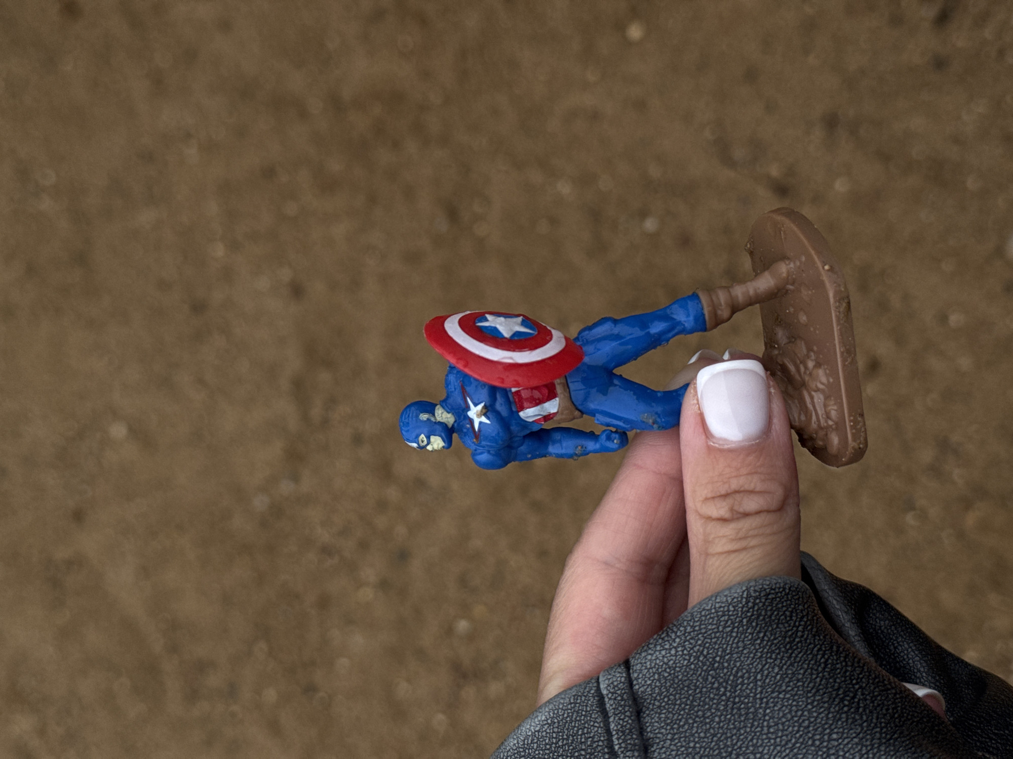In Khimki, Russia, a hand with white-painted fingernails holds a small, blue Captain America toy figure. The figure is detailed with a red and white shield, a star emblem on its chest, and brown boots. The background is a blurred, earthy-brown surface, possibly soil or dirt. The overall scene is an outdoor shot, with the focus on the toy figure being held by a person.