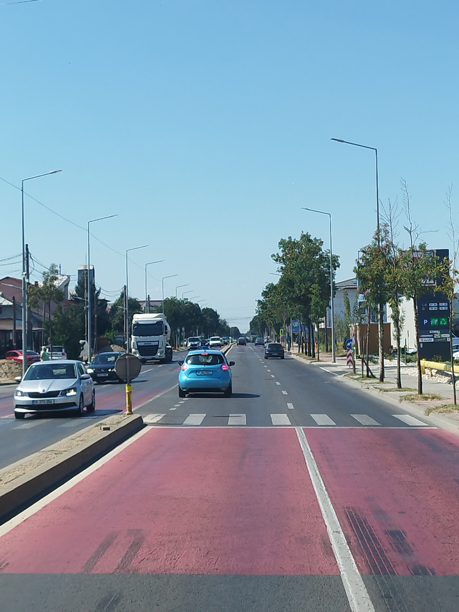 The image shows a street scene in Chitila, Romania, on a clear day. The perspective is from a vehicle looking forward. The road is asphalt with a pedestrian crossing painted in red and white. Several cars are driving on the road, including a silver hatchback in the foreground on the left and a blue hatchback further ahead on the right. A large white truck is also visible in the middle of the road. Streetlights line both sides of the road. Trees and buildings are present on the sides of the street. A sign with prices and logos is visible on the right side of the road. The sky is clear and blue.