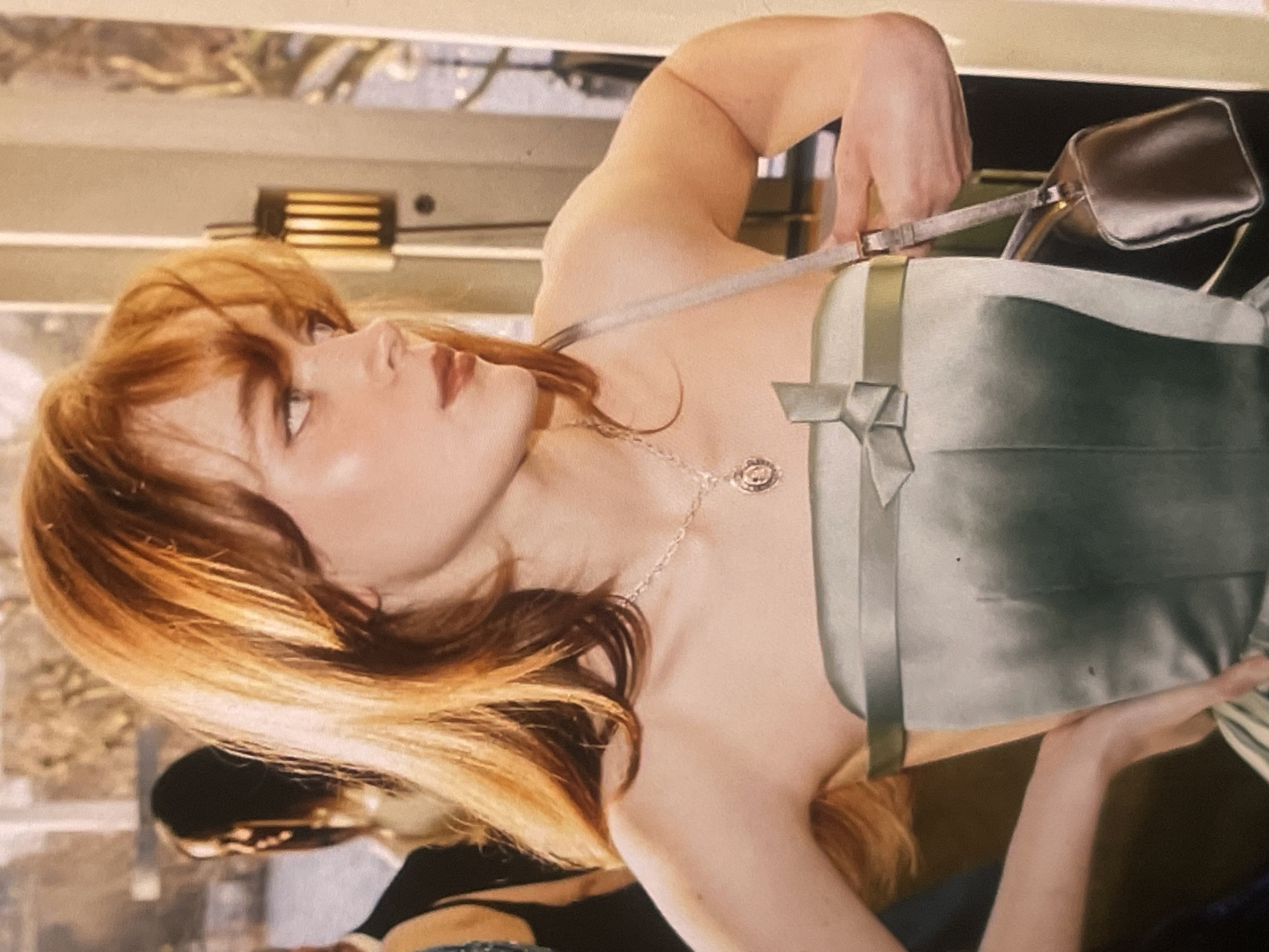 A young woman with long, reddish-blonde hair is captured in an elegant indoor setting. Her head is tilted back, and she gazes upwards and slightly to the left, with a serene and perhaps contemplative expression. Her lips are softly parted. She is wearing a strapless, light sage-green or silvery-green top or dress, made of a lustrous fabric like satin, which features a decorative bow on the left side of the bodice. A delicate silver-toned necklace with a circular pendant adorns her neck.
She is holding a small, metallic silver or champagne-colored handbag with a thin strap, gripping the strap with her right hand. Her skin appears smooth and well-lit.
The background is softly blurred, indicating an interior space. Visible elements include reflections on a surface, a contemporary light fixture with horizontal slats, and a hint of another person in the distance, further obscured by the blur. The overall lighting is soft and bright, suggesting a well-lit indoor environment, possibly a restaurant, cafe, or a stylish building in Poltava, Ukraine. The atmosphere conveys a sense of quiet elegance. There is no visible text in the image.