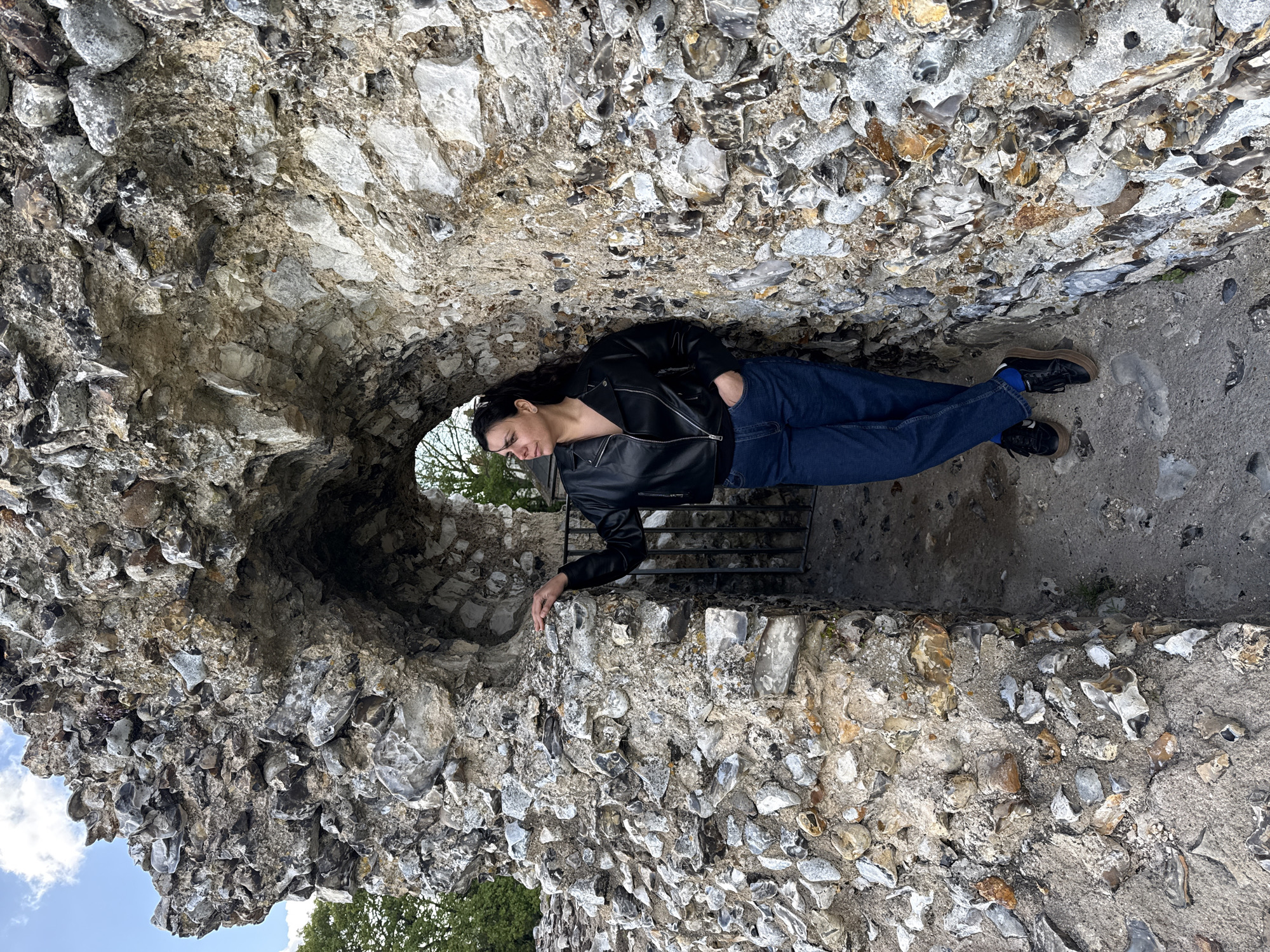 The image, rotated 90 degrees clockwise, displays a person lying down within a rustic stone archway or tunnel. The archway is constructed from irregular, rough-hewn stones, predominantly grey, white, and brown, resembling flint, set in mortar. The texture of the stone work is highly varied and uneven.
The subject is a person with dark hair, dressed in a black leather-style jacket over a dark, possibly unbuttoned top, blue jeans, and dark shoes with light brown soles. Blue socks are visible above the shoes. One hand is casually placed in a jeans pocket, while the other rests gently on the rough stone wall of the archway. The person's gaze is directed slightly upwards towards the opening, exhibiting a calm expression with a subtle smile.
Through the archway's far opening, a bright blue sky with patches of white clouds is visible, along with green foliage from trees, indicating a clear, daytime setting. A dark metal gate or grille is partially visible within the deeper shadows of the archway, behind the person's head. The ground or floor inside the archway, beneath the person's feet, appears to be made of concrete or packed earth interspersed with small stones. The distinct flint architecture suggests an ancient or historical structure, characteristic of sites found in the King's Lynn and West Norfolk area of the United Kingdom. No text is visible in the image.