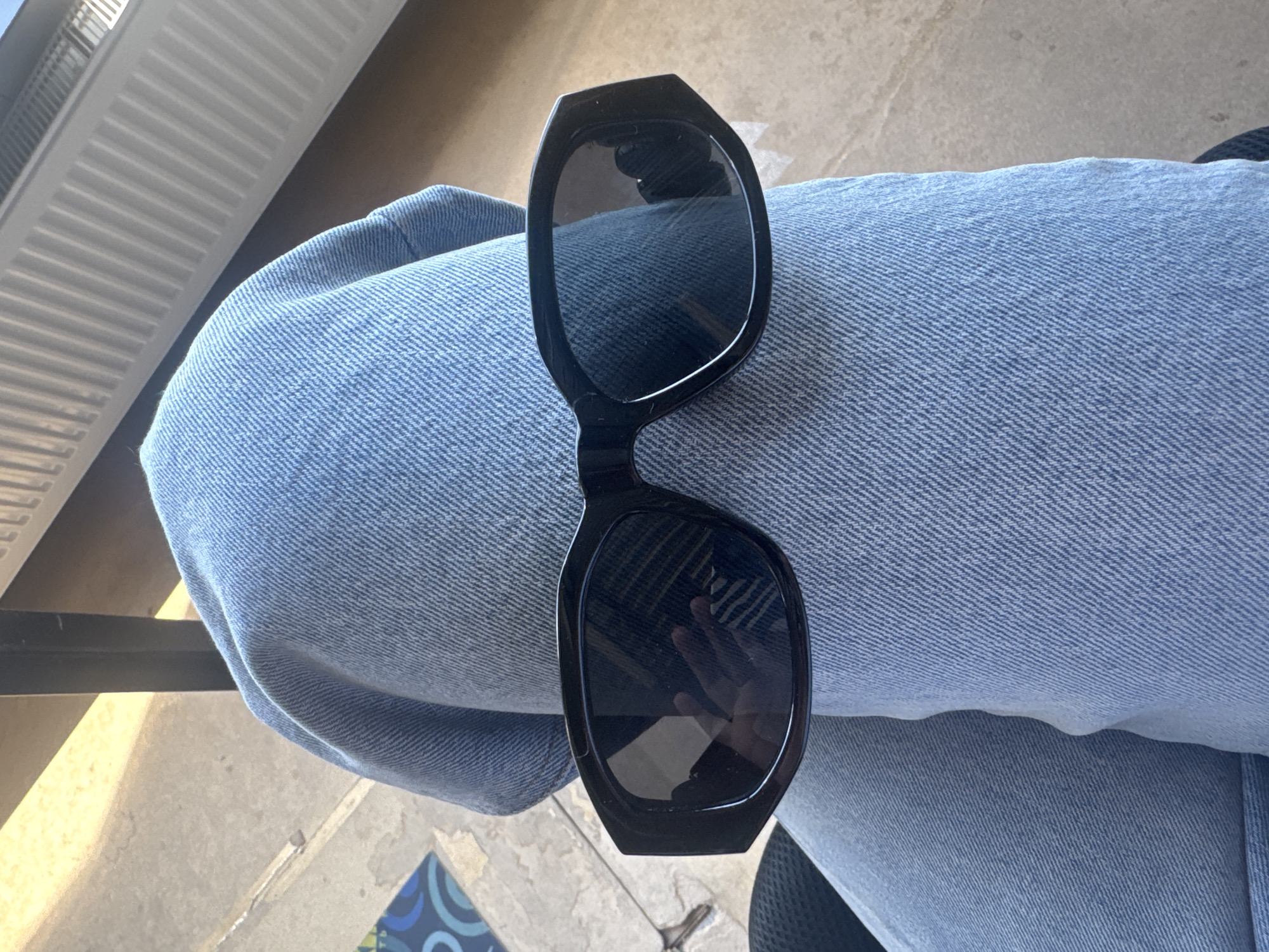A pair of black sunglasses sits atop a person's lap, covered in blue denim jeans. In the background, a radiator is visible. A partially visible chair and a patterned object, perhaps a cushion, can be seen near the bottom edge of the frame. The location is Chaikovsky, Russia.