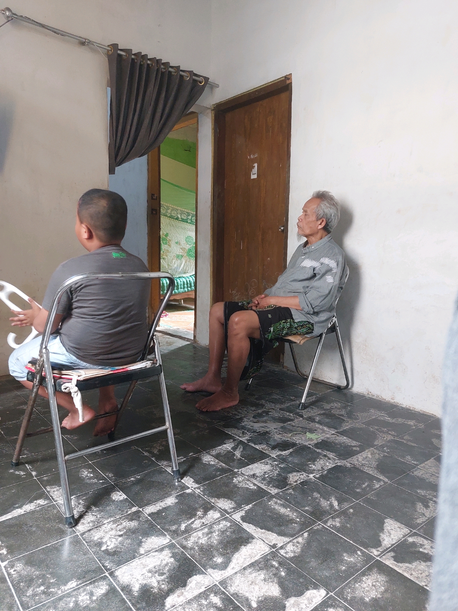 The image depicts two individuals in a room, likely in Sarangan, Indonesia. In the foreground, a young boy with short hair is seated on a folding metal chair. He is facing away from the camera, wearing a grey t-shirt and light-colored shorts. His left hand is holding a white object with multiple circular openings.
Further back and to the right, an older man with grey hair is seated on a similar folding metal chair. He is facing to the left, looking towards the boy and a doorway. He is wearing a grey collared shirt and dark shorts, with a patterned cloth draped over his right leg. His hands are clasped in his lap.
The room has white walls and a dark tiled floor with visible wear and tear. To the left, a dark brown curtain is partially drawn, revealing a glimpse of an adjacent room with green patterned bedding. A wooden door is visible behind the older man, and above it, a label with text is present. The overall impression is of a simple, indoor domestic setting.