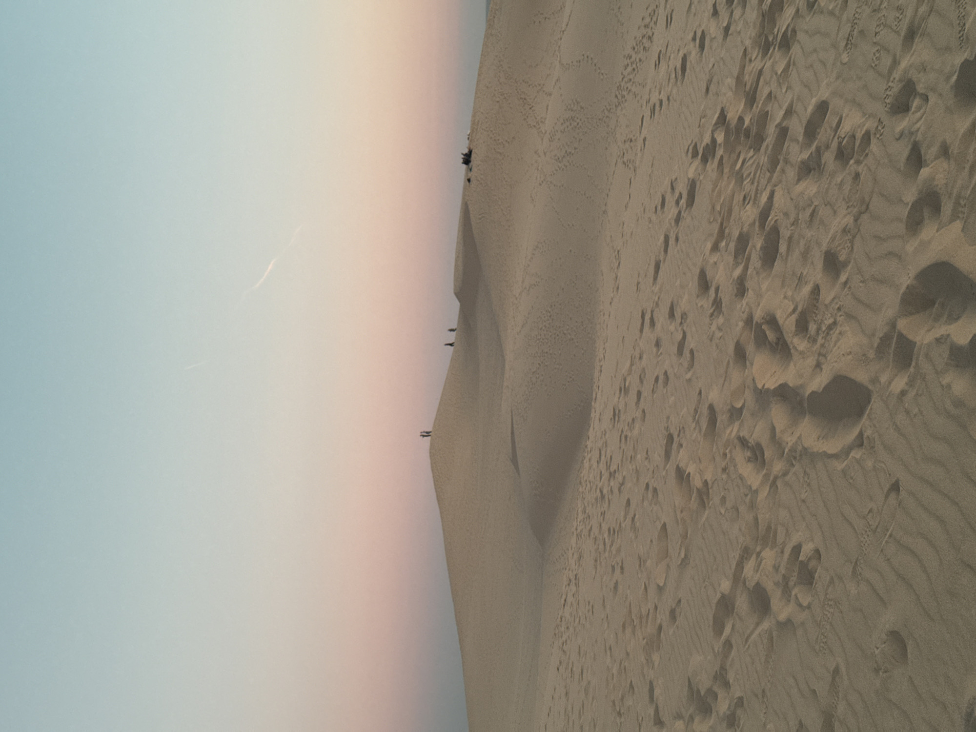 The image depicts a panoramic view of the Dune of Pilat, located in La Teste-de-Buch, France. The sand dunes stretch across the frame, with footprints visible on the sandy surface in the foreground. In the middle ground, several individuals are scattered on the crest of a dune, some standing and others appearing to be resting. The sky above transitions from a pale blue at the top to a soft pink and orange hue near the horizon, suggesting it is either sunrise or sunset. The overall scene conveys a sense of vastness and natural beauty.