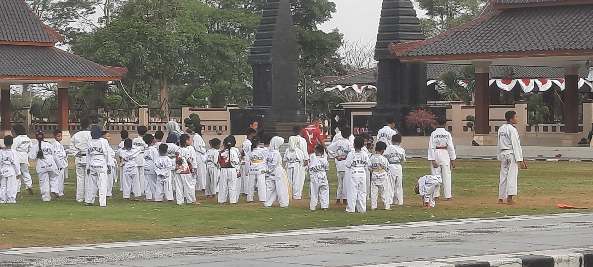 A group of young people, primarily children, are gathered on a grassy outdoor area in what appears to be a public space. They are dressed in white taekwondo uniforms with varying belt colors, indicating different levels of proficiency. The children are facing forward, and some are looking towards an unseen point of interest. In the background, there are traditional Indonesian architectural structures, including a building with a dark, tiered roof and decorative pillars, as well as a large, dark, monument-like structure with layered tiers. Red and white decorative banners are visible on the building in the background. The ground is covered in green grass, and a paved path or road is in the foreground. The lighting suggests it is daytime, with an overcast sky. The overall scene suggests an organized event or training session for young taekwondo practitioners. The presence of specific architectural styles suggests a location within Indonesia, and local knowledge would be required to pinpoint the exact city and neighborhood within Kanigoro.