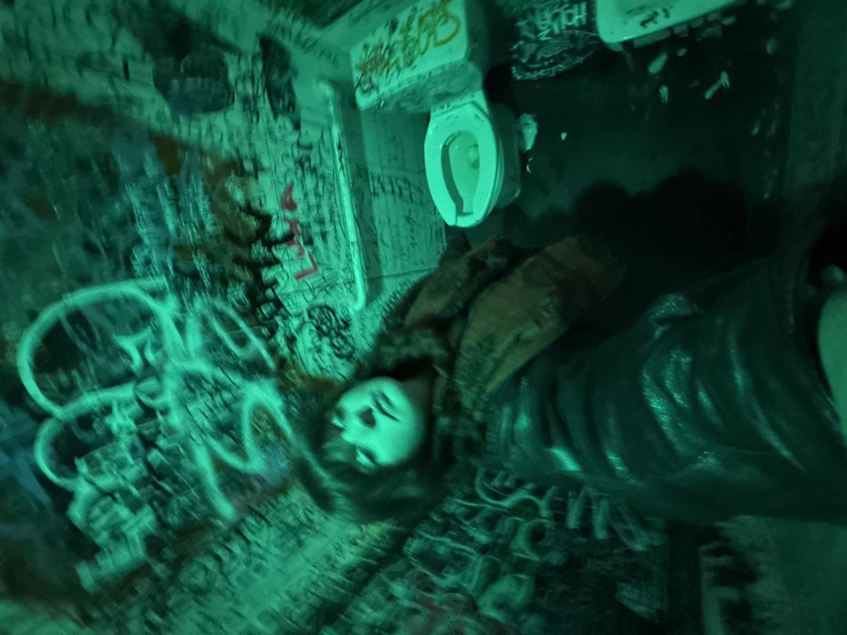 A person in a dark, graffiti-covered room looks downward, with a toilet mounted above. Walls and surfaces are covered in dense, colorful graffiti. The person, wearing dark clothing, is visible from the waist up, and is located at the center of the frame. A green hue dominates the image, casting an eerie glow on the scene. The overall composition feels claustrophobic and intense.
