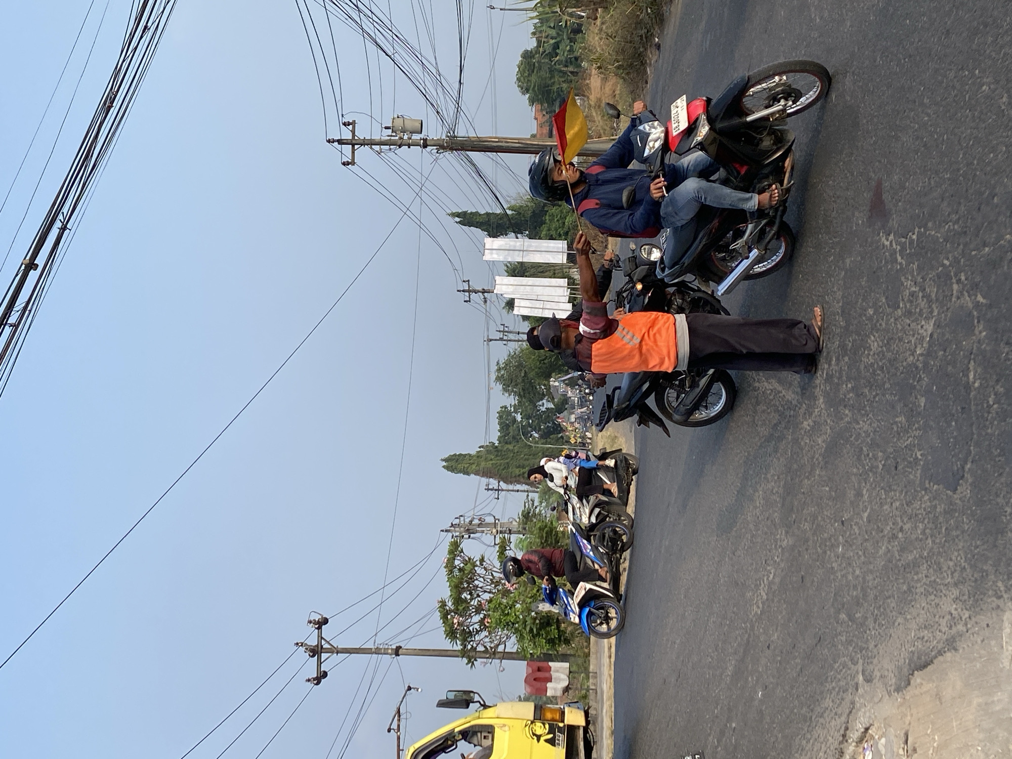 In Kasihan, Indonesia, a traffic director wearing an orange vest and dark pants stands on the right side of a two-lane road, gesturing with his right hand. He is facing left, towards a motorcyclist wearing a blue jacket and helmet, who is riding a black motorcycle with a red front fender and license plate. The motorcyclist holds a red and yellow flag in his right hand. Behind this motorcyclist, another person is seated on a black motorcycle. To the left of the traffic director, another motorcycle with a rider is also visible. Further to the left, a yellow truck is parked on the side of the road. The road is paved with asphalt, and utility poles with numerous wires are visible in the background. The sky is clear and blue, with the sun casting shadows on the road. Trees and vegetation line the sides of the road.