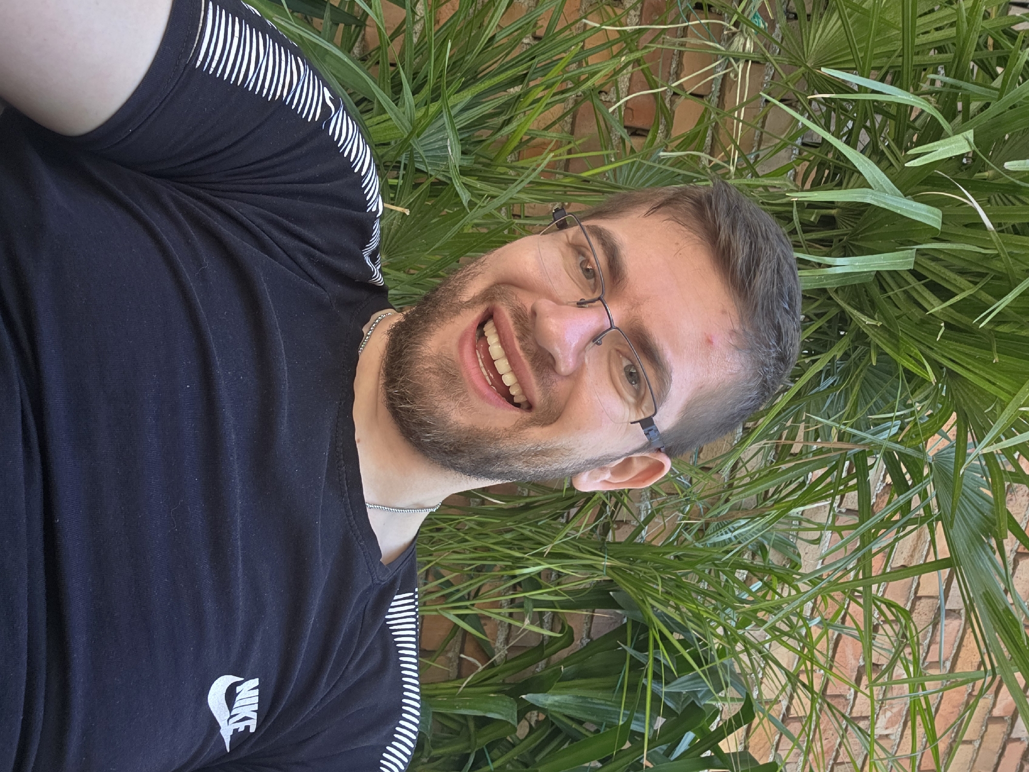 A man with short brown hair, a beard, and glasses is smiling and looking at the camera. He is wearing a black t-shirt with white stripes on the shoulders and a white Nike logo on the front. He is wearing a silver chain around his neck. The background includes green palm-like plants and a brick wall. The location is not explicitly evident to be Yagodovo, Bulgaria.