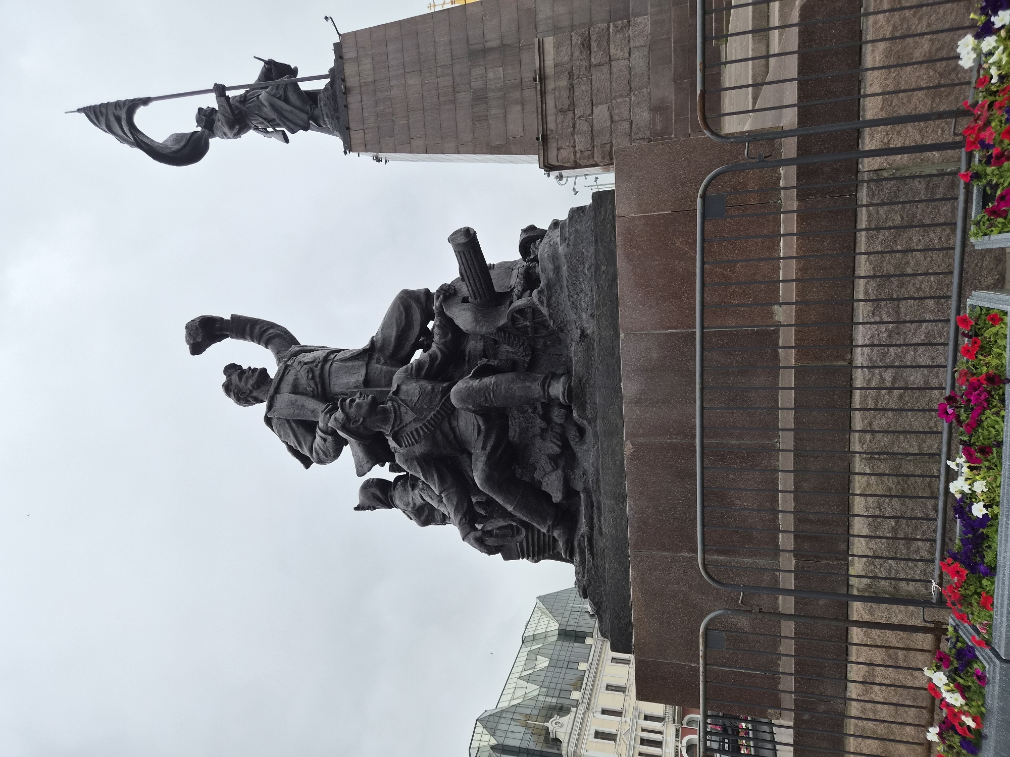 This image depicts the monument to the fighters for Soviet power in the Far East, located in Vladivostok, Russia. The monument is a large bronze sculpture featuring several figures, including soldiers and a civilian, in a dynamic composition. One figure stands tall, raising a fist, while another is seated, holding a rifle. A cannon is also visible within the sculpture. In the background, a slender obelisk topped with a statue holding a flag stands prominently against a cloudy sky. To the left of the monument, a modern building with a glass-paneled roof and arched windows can be seen. In the foreground, temporary metal barriers are erected, and flower planters filled with red, white, and purple flowers are placed in front of them. The perspective of the photo is from a low angle, looking up at the monument.