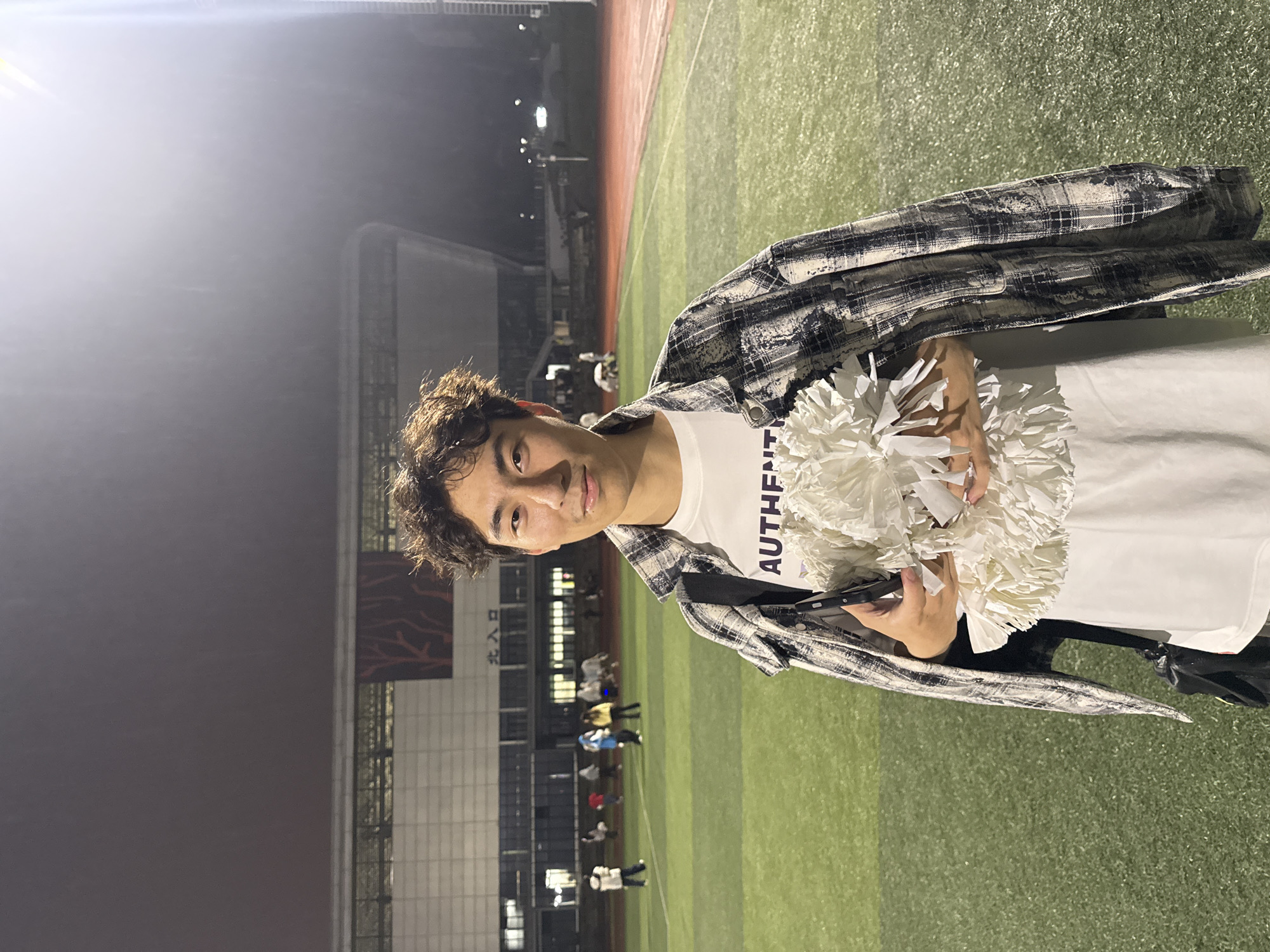 A young man stands on a sports field, likely a track and field or soccer field, at night. He is holding white pom-poms, suggesting he might be a cheerleader or involved in a similar activity. He is wearing a white t-shirt with "AUTHENTI" visible and a black and white checkered jacket. In the background, there are other people on the field, and stadium seating is visible. The image seems to have been taken near the entrance to the stadium, as indicated by Chinese characters. The location is Jiangjin, China.