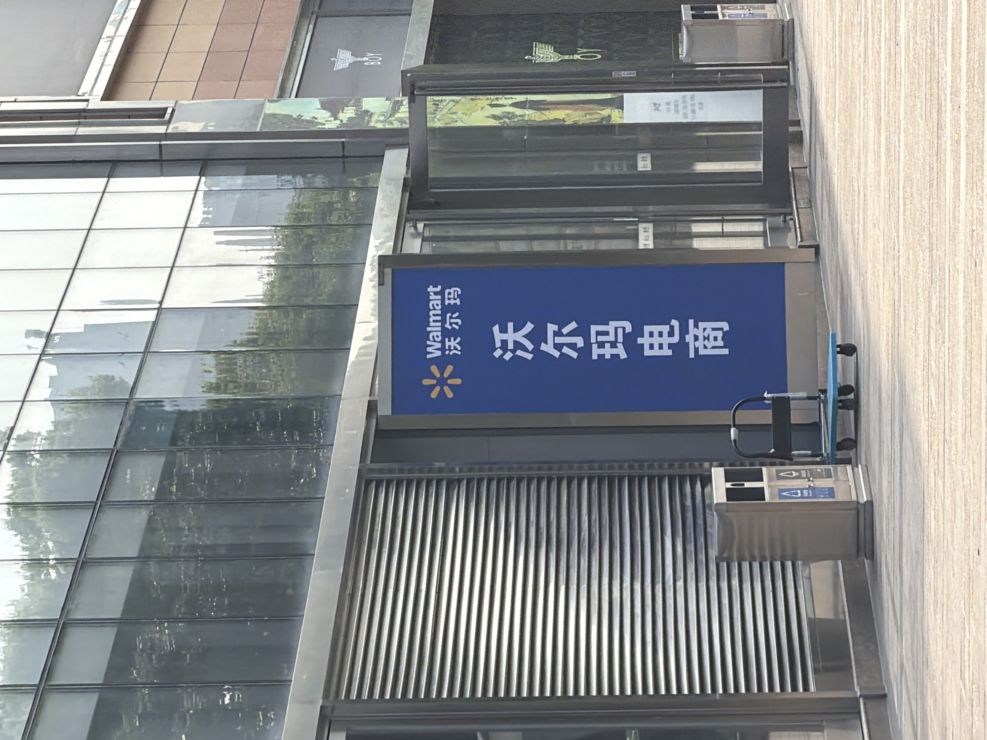 The image shows a modern building in Yuhuatai District, Nanjing, China. A large Walmart e-commerce sign is visible, mounted on the side of the building. The sign is blue with the Walmart logo and Chinese characters. The building has glass panels reflecting the surroundings. A metal waste disposal unit is attached to the building near the sign. The sidewalk is light-colored and runs along the building. No people are visible in the immediate scene.