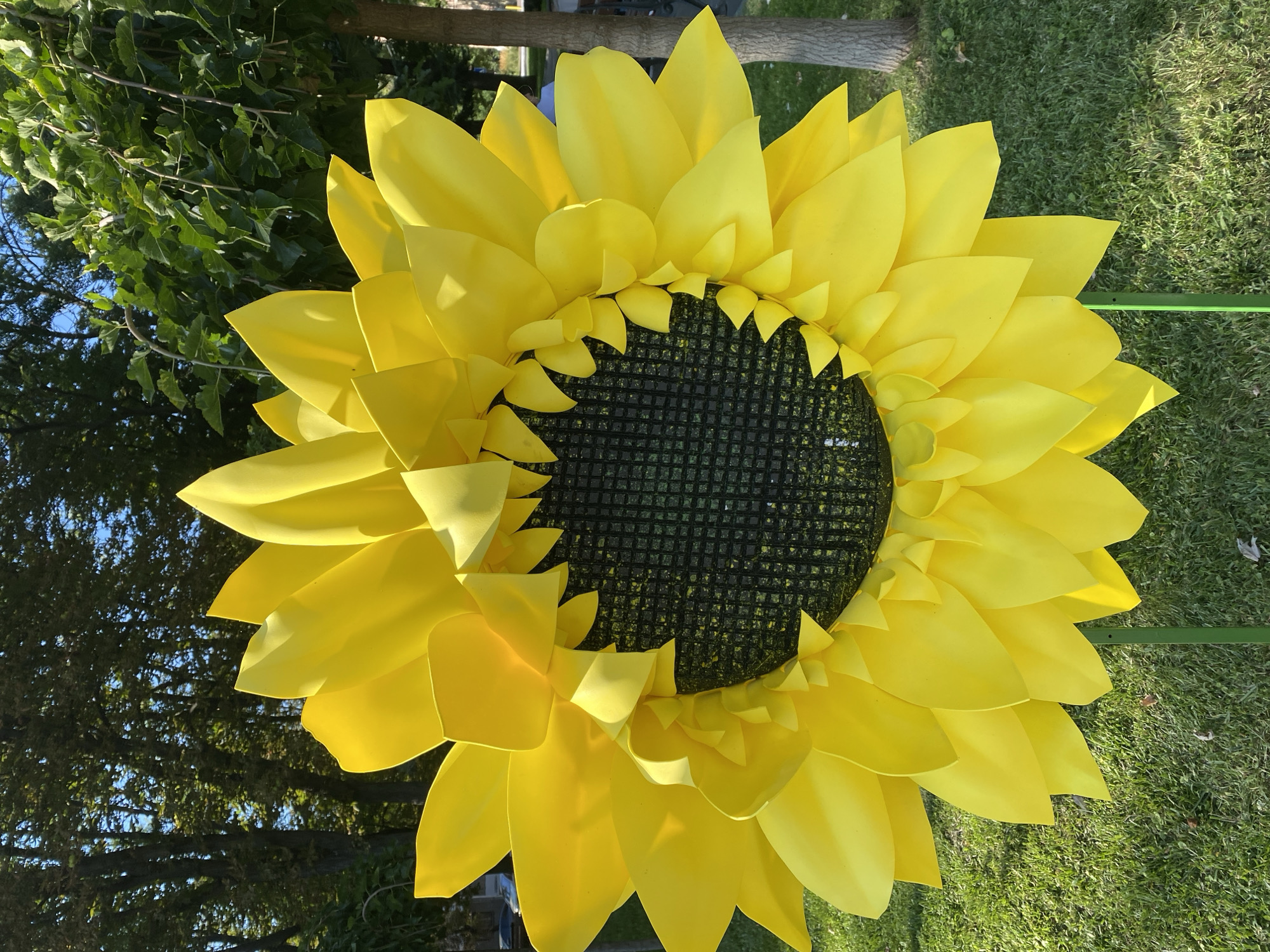 A large, artificial sunflower decoration stands in a grassy area in Kremenchuk, Ukraine. The sunflower has bright yellow petals and a dark, checkered center. It is supported by two green poles. Behind the sunflower, trees with green leaves and a blue sky are visible. The ground is covered in green grass with some fallen leaves.