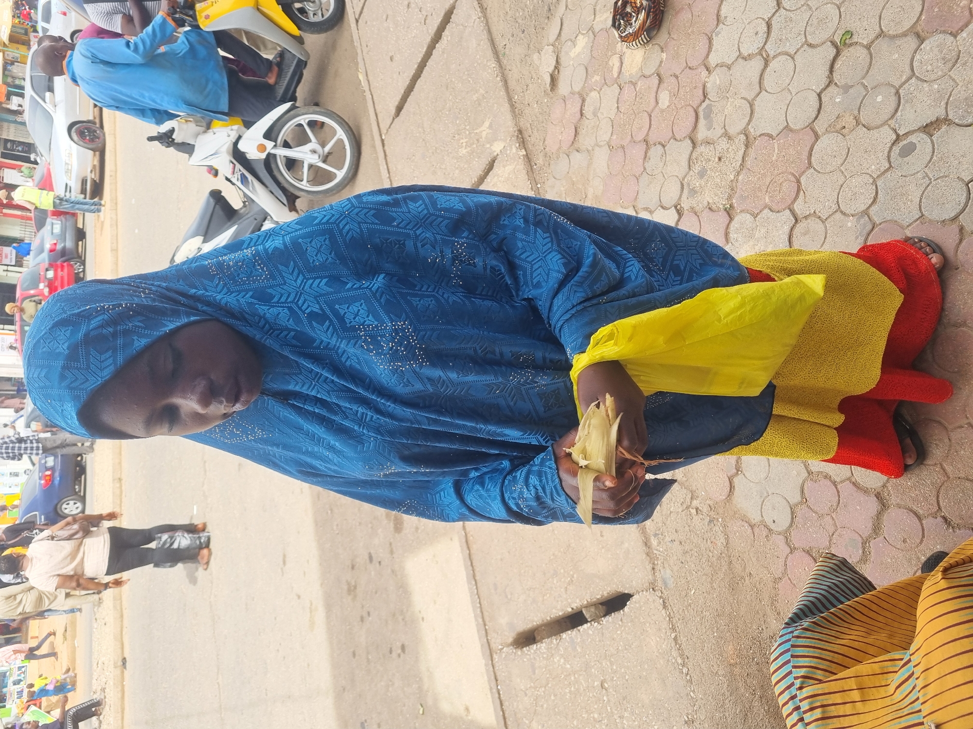 A woman wearing a blue patterned hijab, a yellow scarf, and a colorful yellow-red skirt sits on the sidewalk in Jos, Nigeria. She is holding food wrapped in a husk. The sidewalk is made of hexagonal concrete pavers, and a drainage grate is visible in the foreground. In the background, there are vehicles, including a scooter, and several pedestrians.